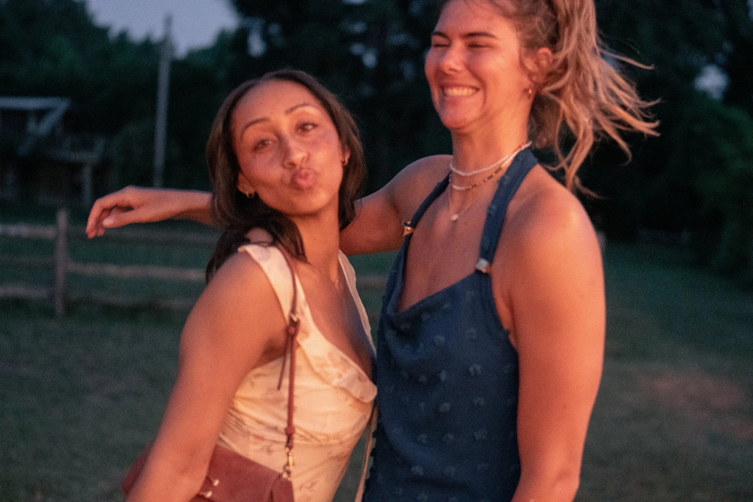 Two women smiling and hugging outdoors during sunset, with trees and a basketball hoop in the background.