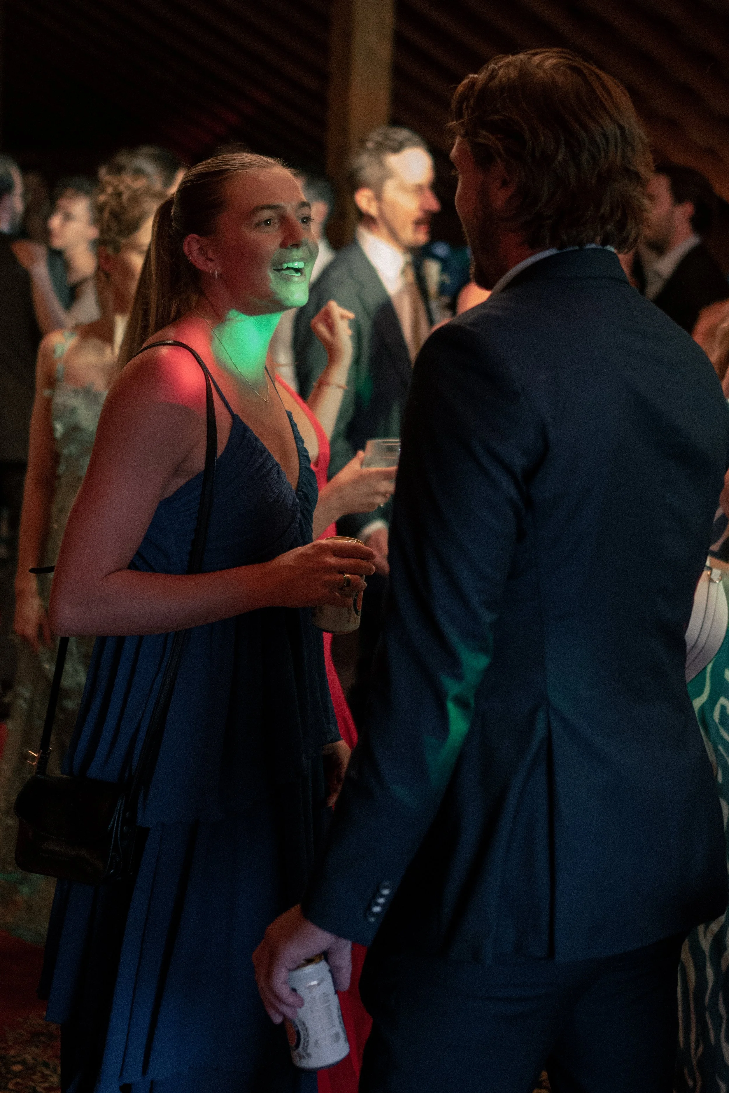 A woman and a man are having a conversation at a social event. The woman is wearing a dark blue dress with thin straps, has her hair pulled back, and is holding a drink and a can of soda. The man, seen from the back, has shoulder-length hair and is d