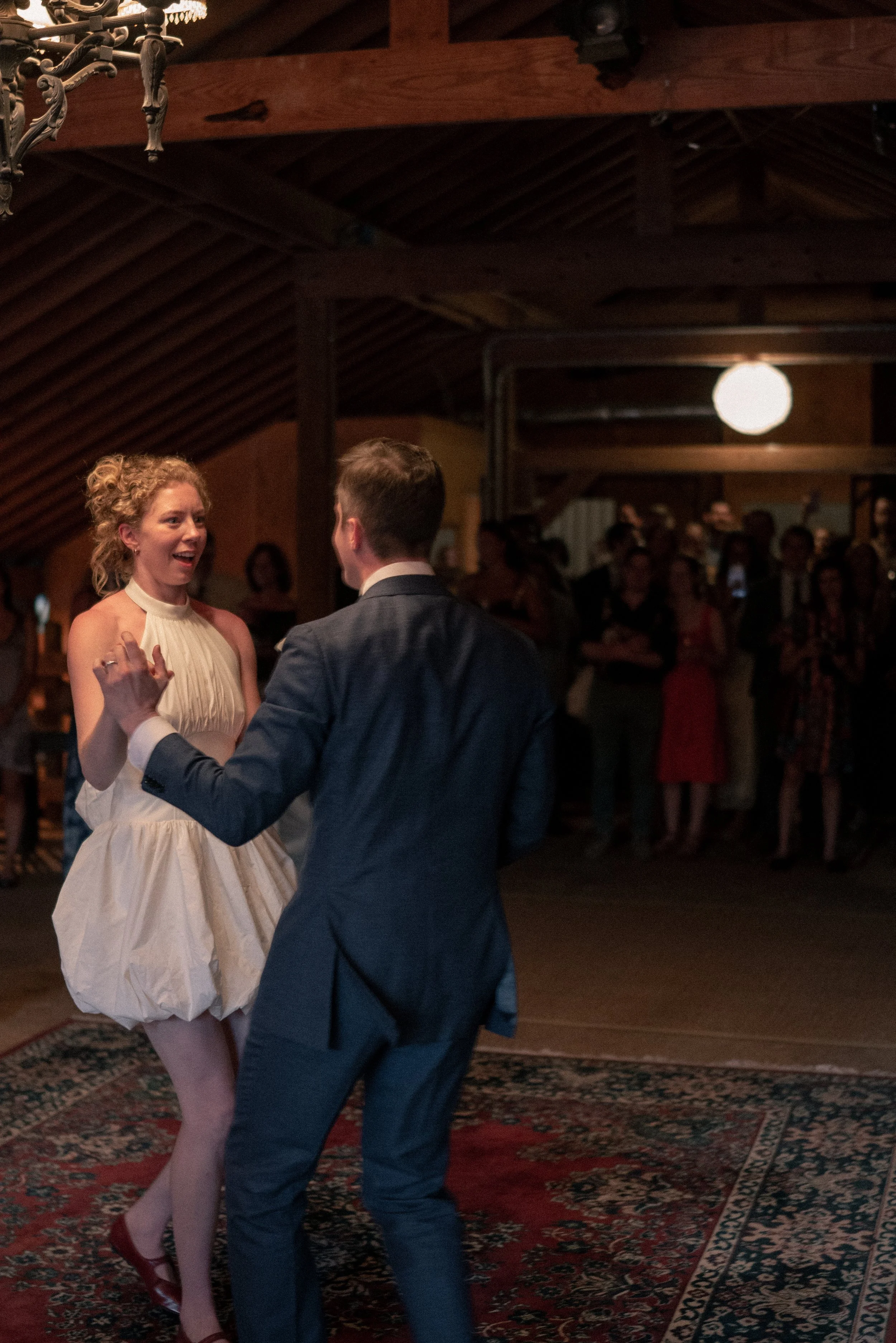 A woman in a white dress and a man in a suit dancing together at an indoor event with a crowd in the background.