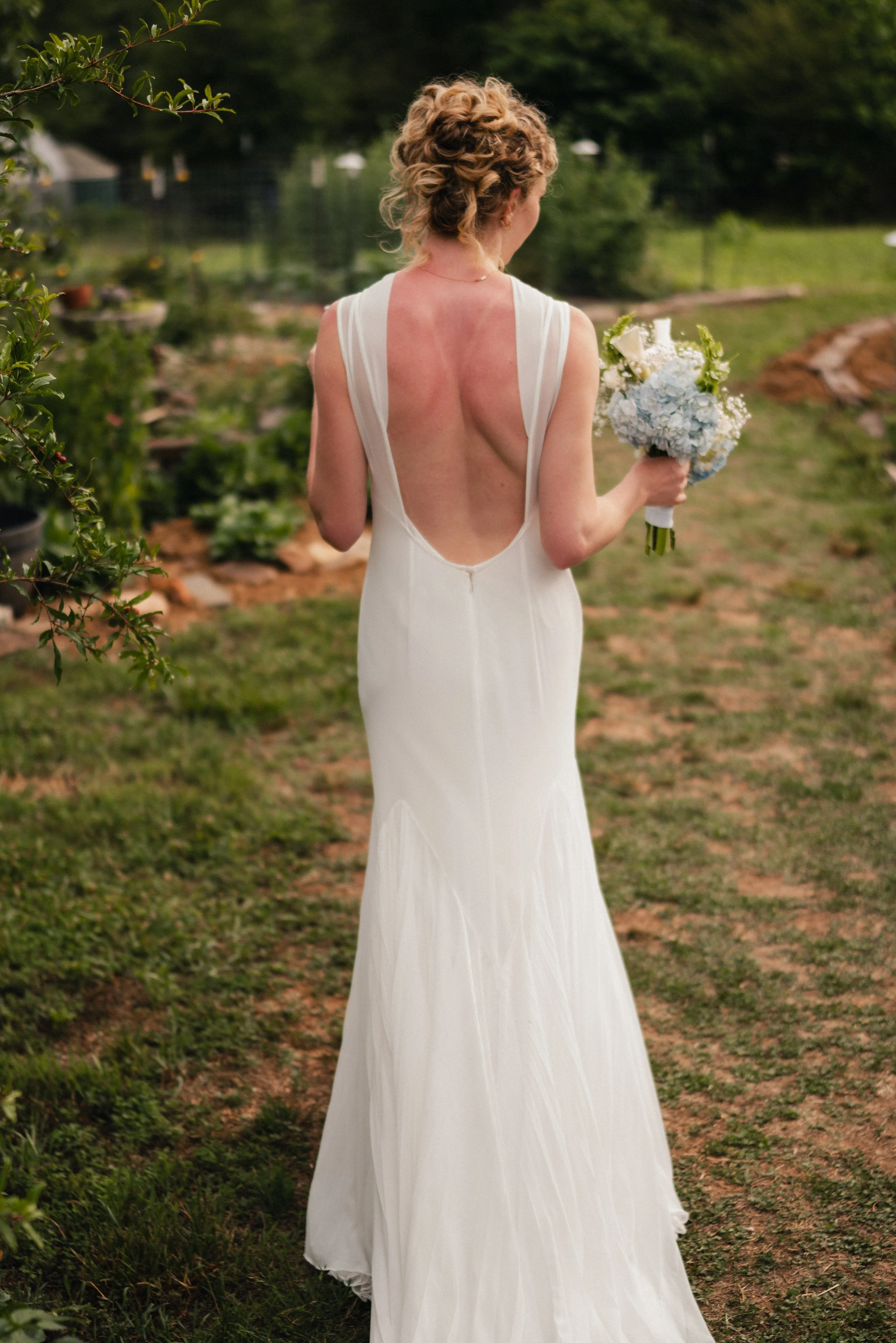 A woman in a white dress with an open back holding a bouquet of white and light blue flowers, standing outdoors on a grassy area with trees and a pond in the background.