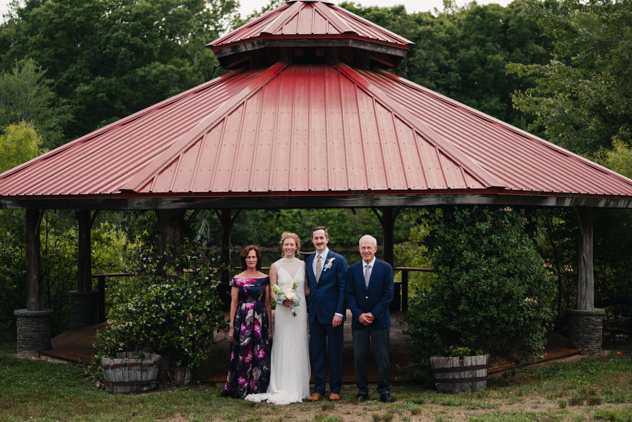 A wedding couple standing outdoors under a red metal-roofed gazebo, with three other people surrounding them, in front of green trees and shrubbery.