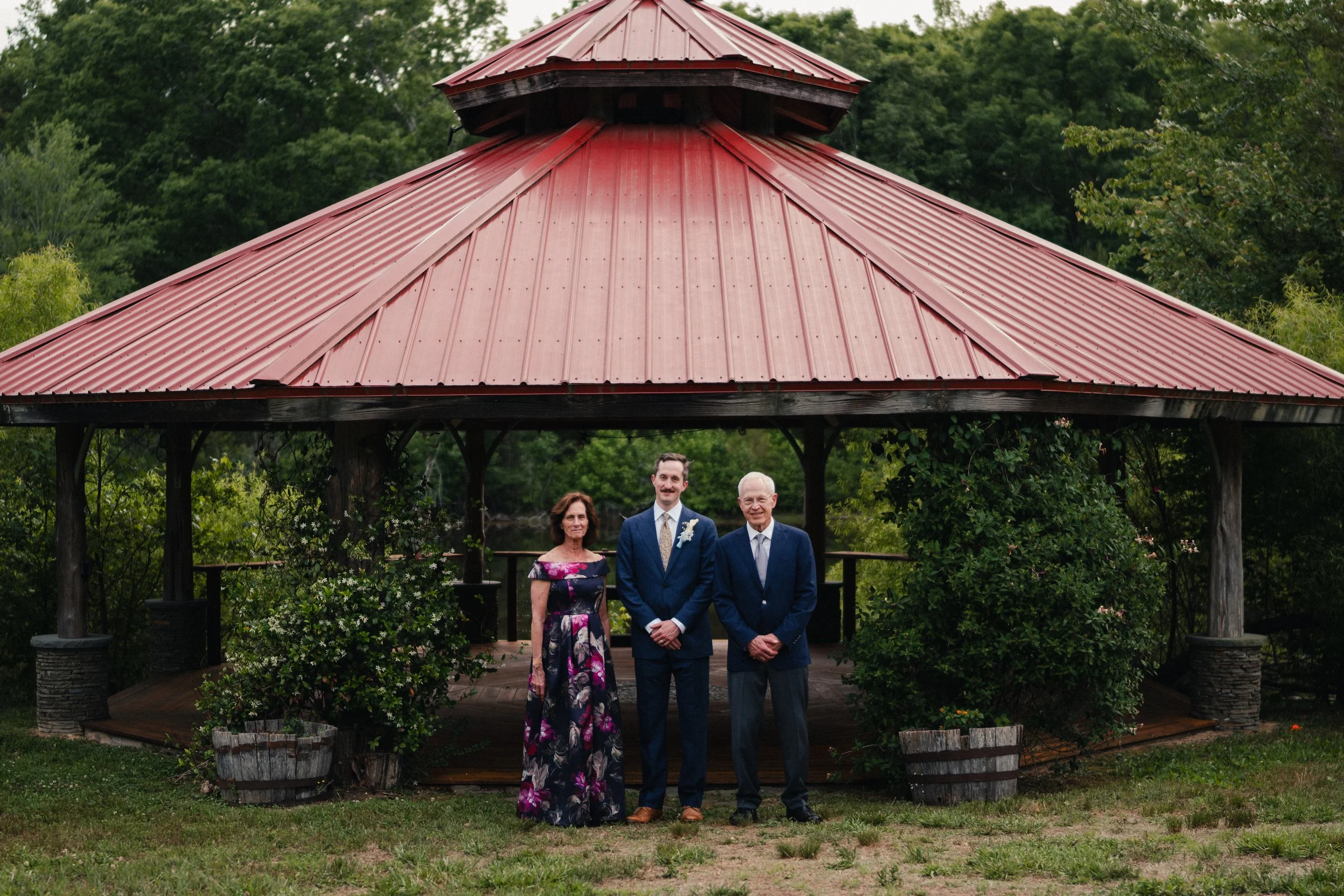 Three people standing together in front of a gazebo with a red metal roof, surrounded by greenery and trees.