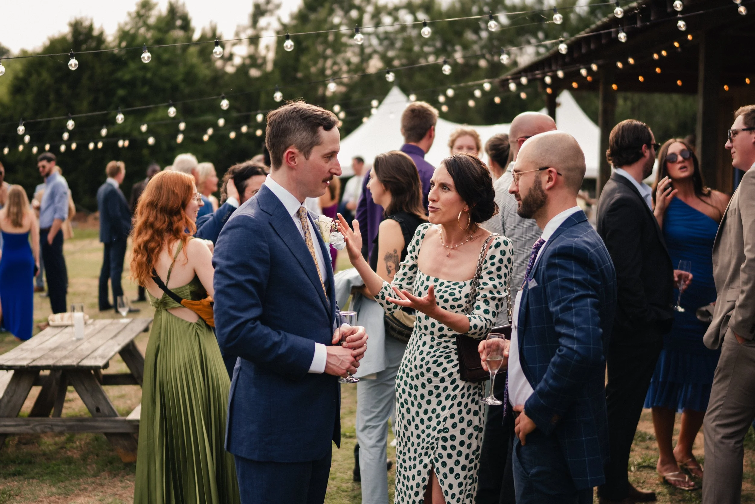 People socializing at an outdoor event during the evening, wearing formal attire, with string lights overhead.