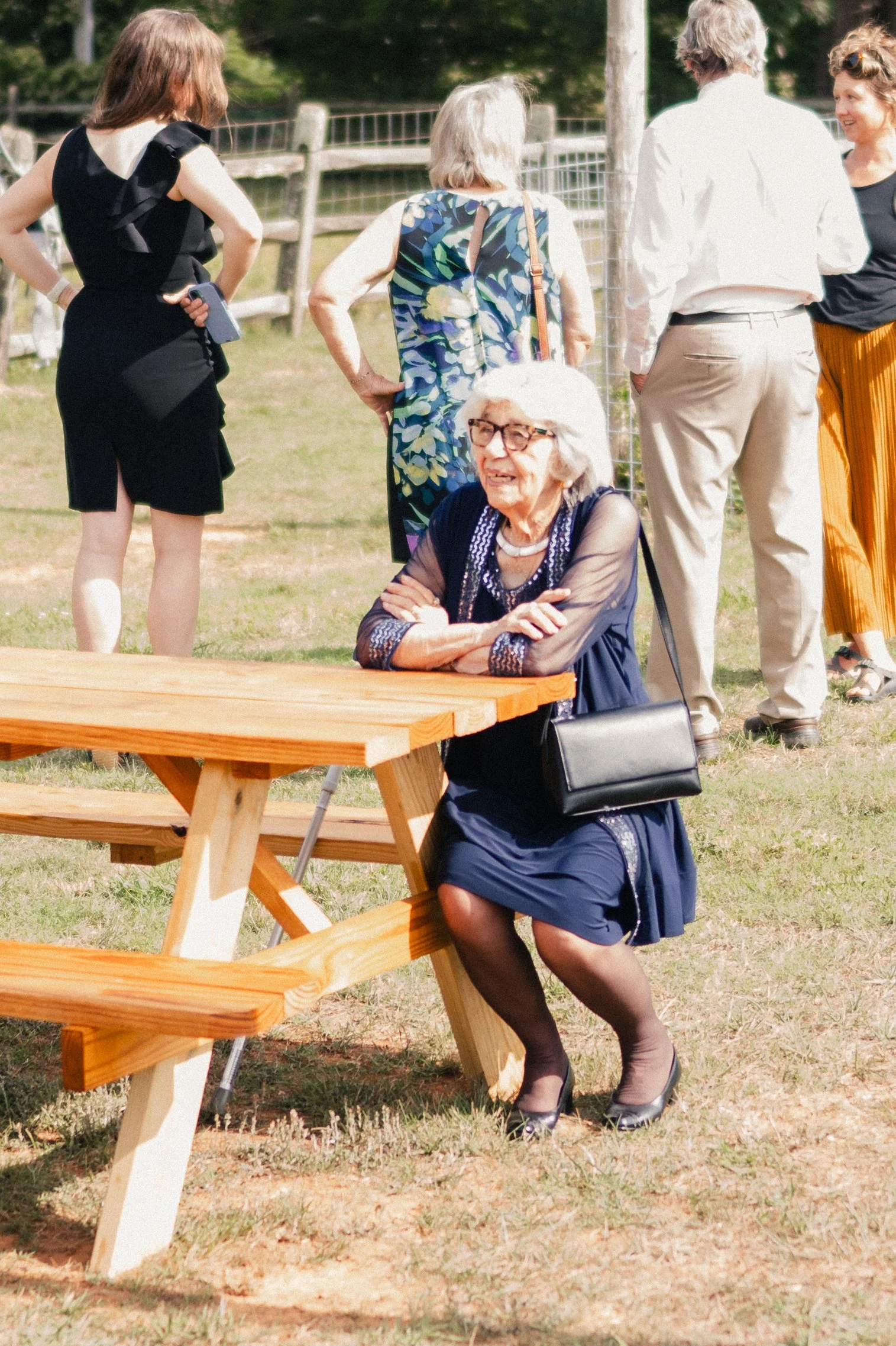 A group of five women socializing outdoors near a wooden picnic table, with green trees and a fence in the background.