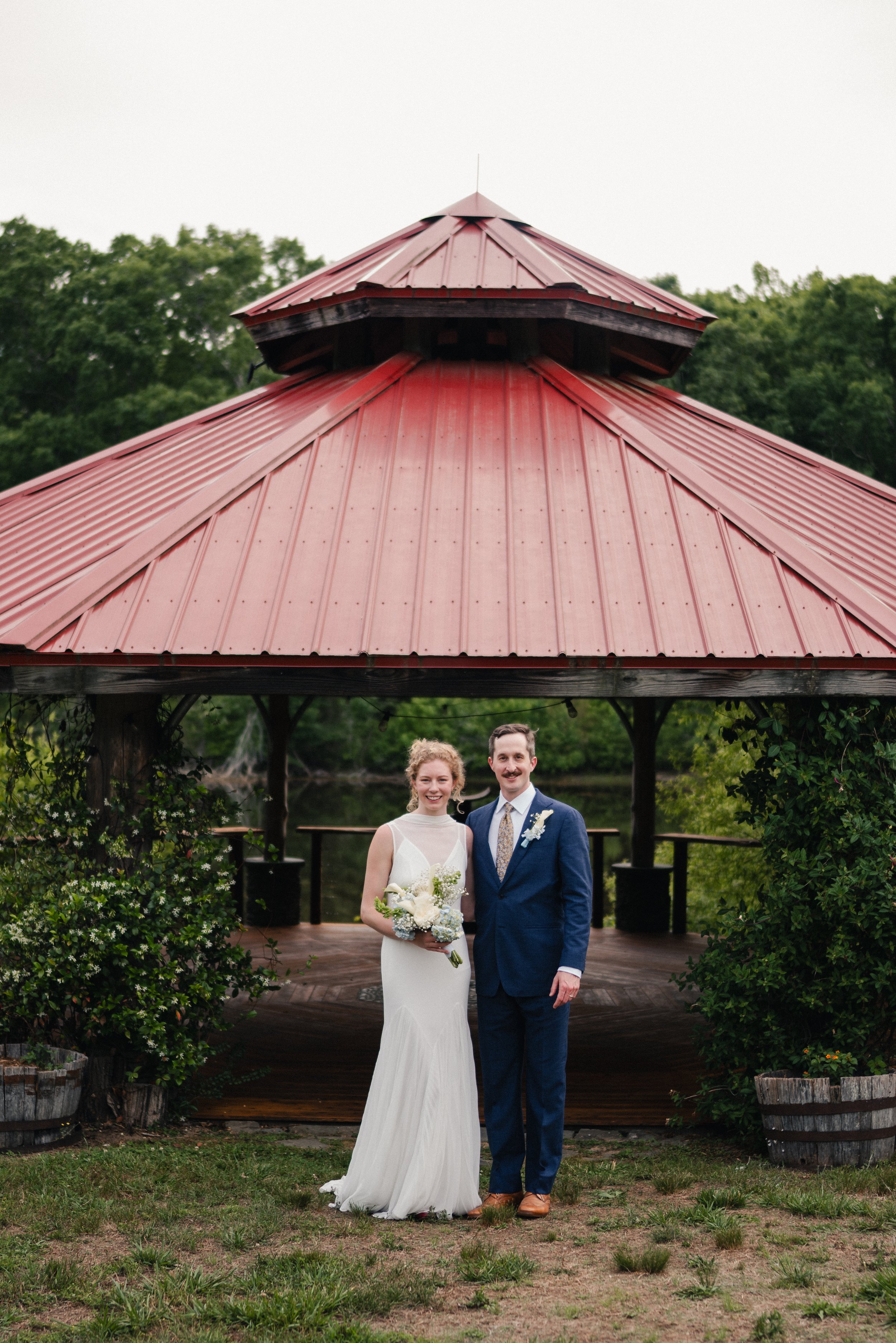 A bride and groom standing together outdoors underneath a red metal roof gazebo, surrounded by greenery and wooden barrels, with a river in the background.