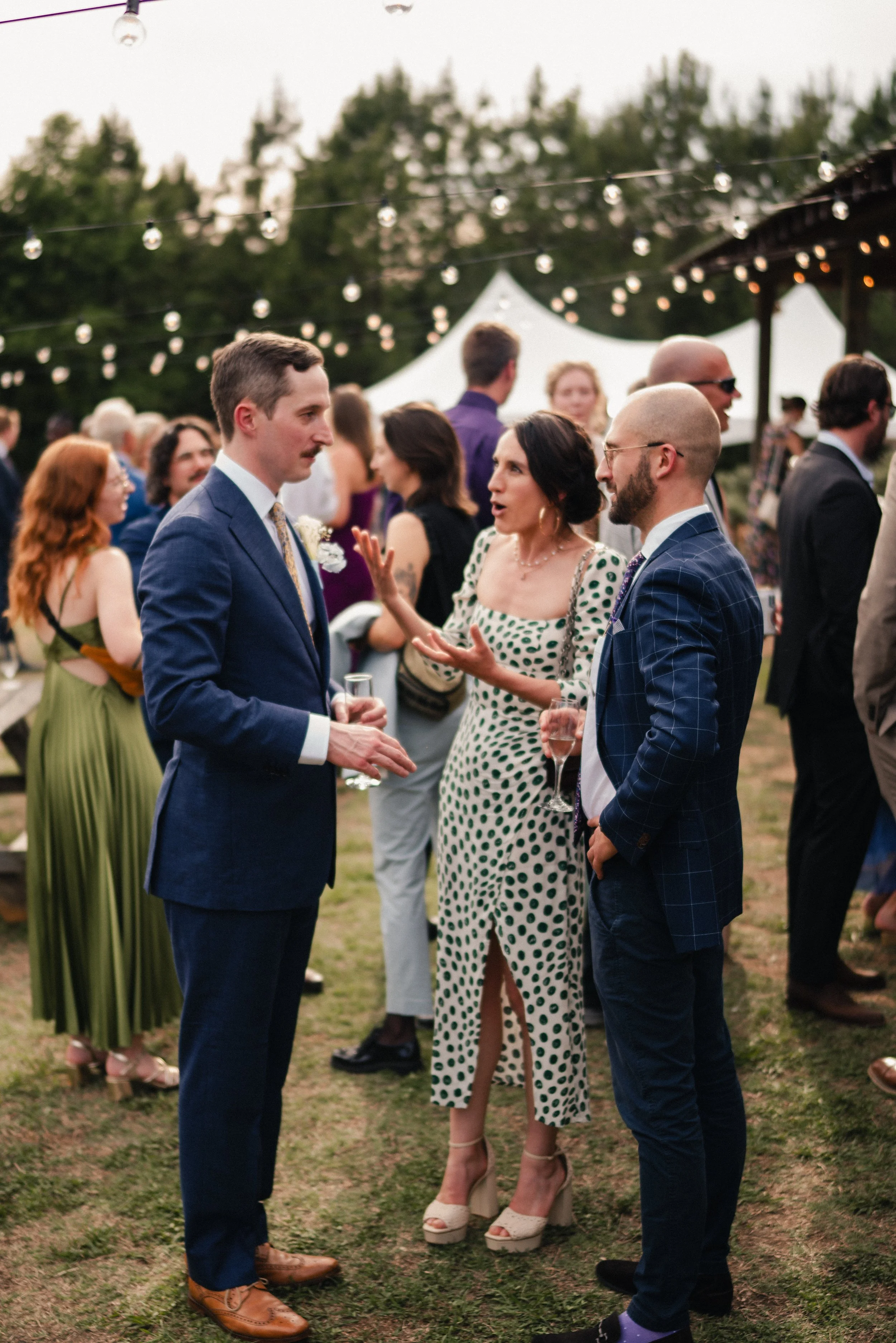 Guests at an outdoor party engage in conversation, with a man in a navy suit and a woman in a polka dot dress speaking animatedly, while other guests mingle in the background under string lights and a white tent.