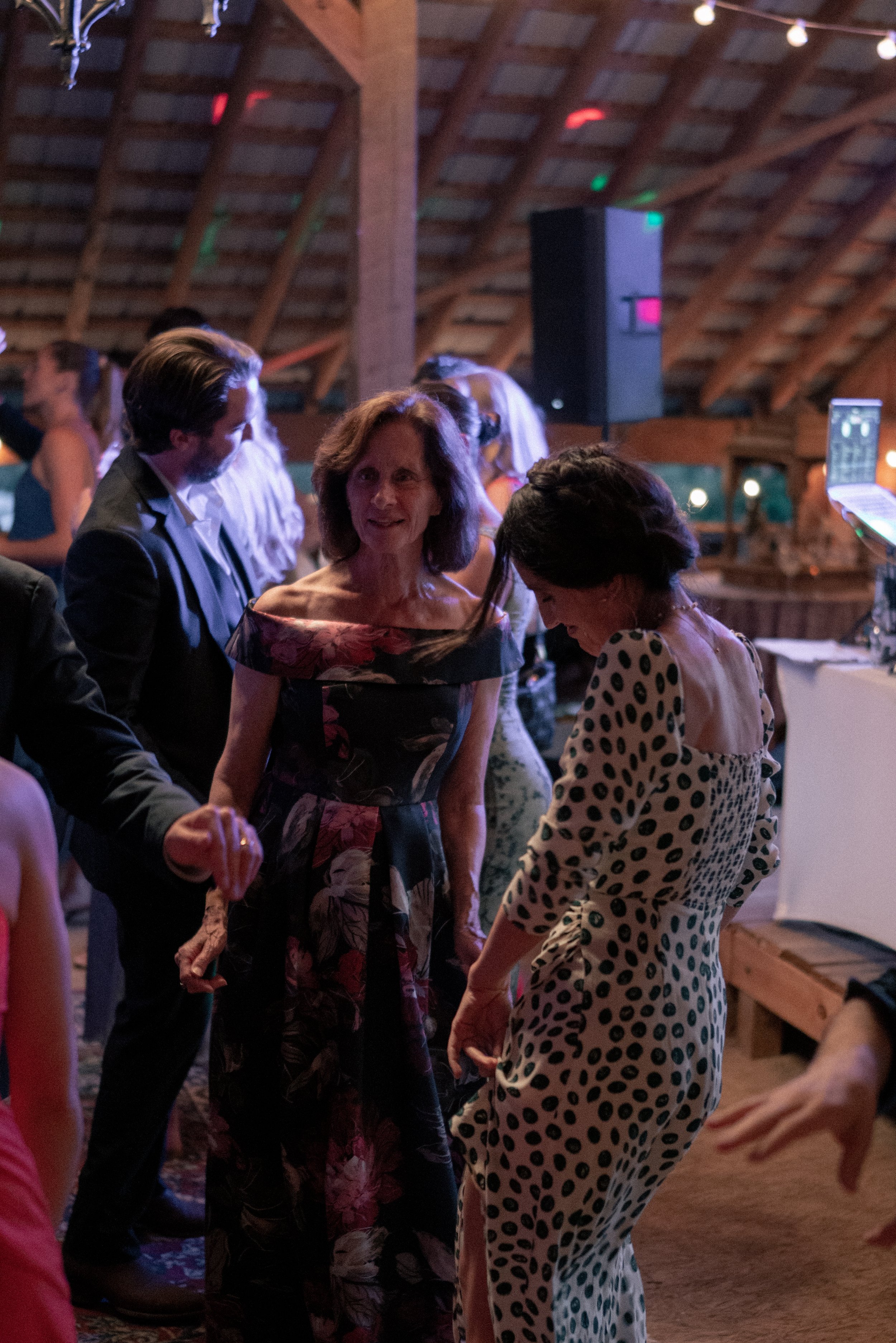 People dancing and socializing at a nighttime event inside a wooden venue with a high, sloped ceiling and string lights.