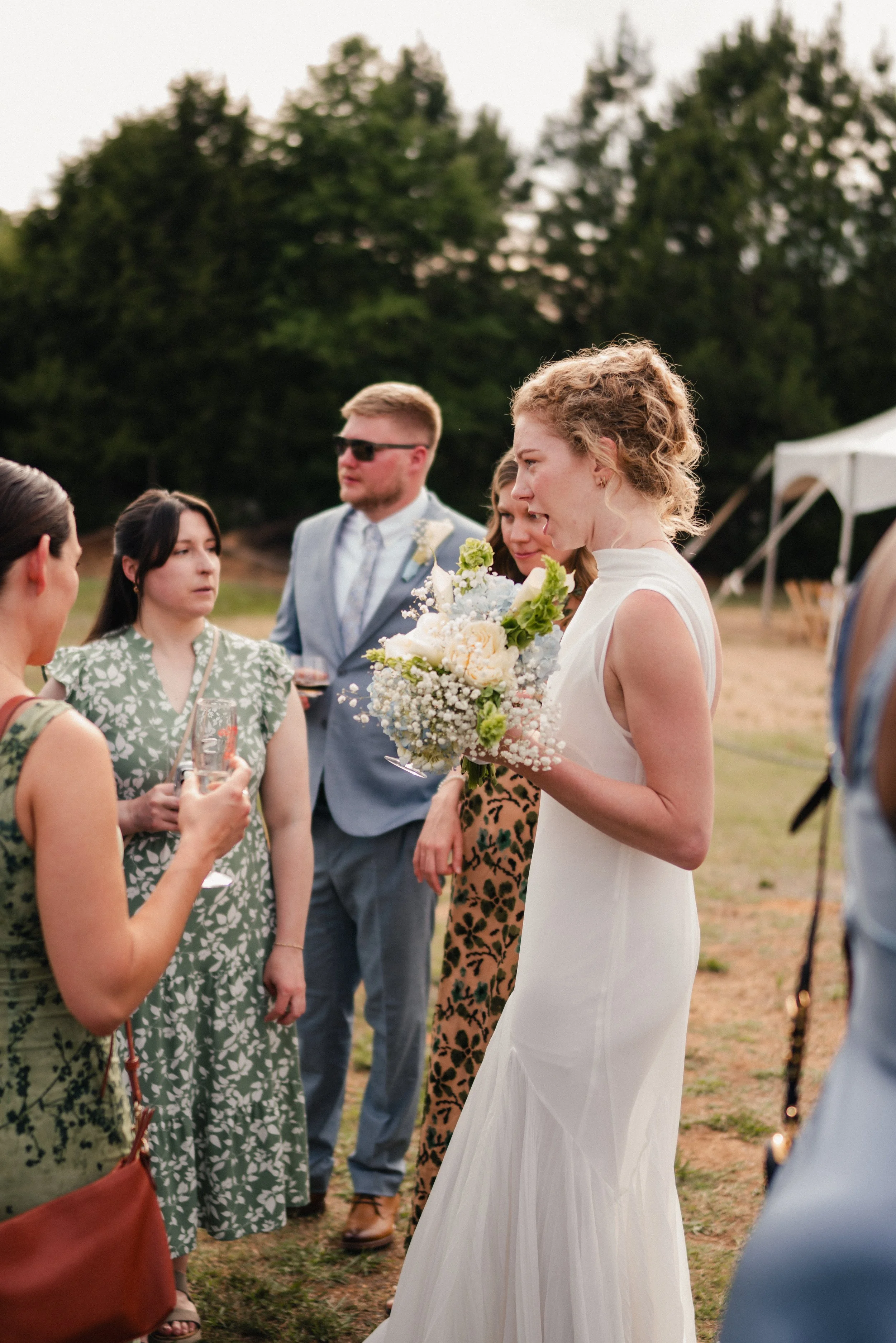 A woman in a white dress holding a bouquet of white and green flowers, speaking to a group of people outdoors during sunset, with trees in the background.