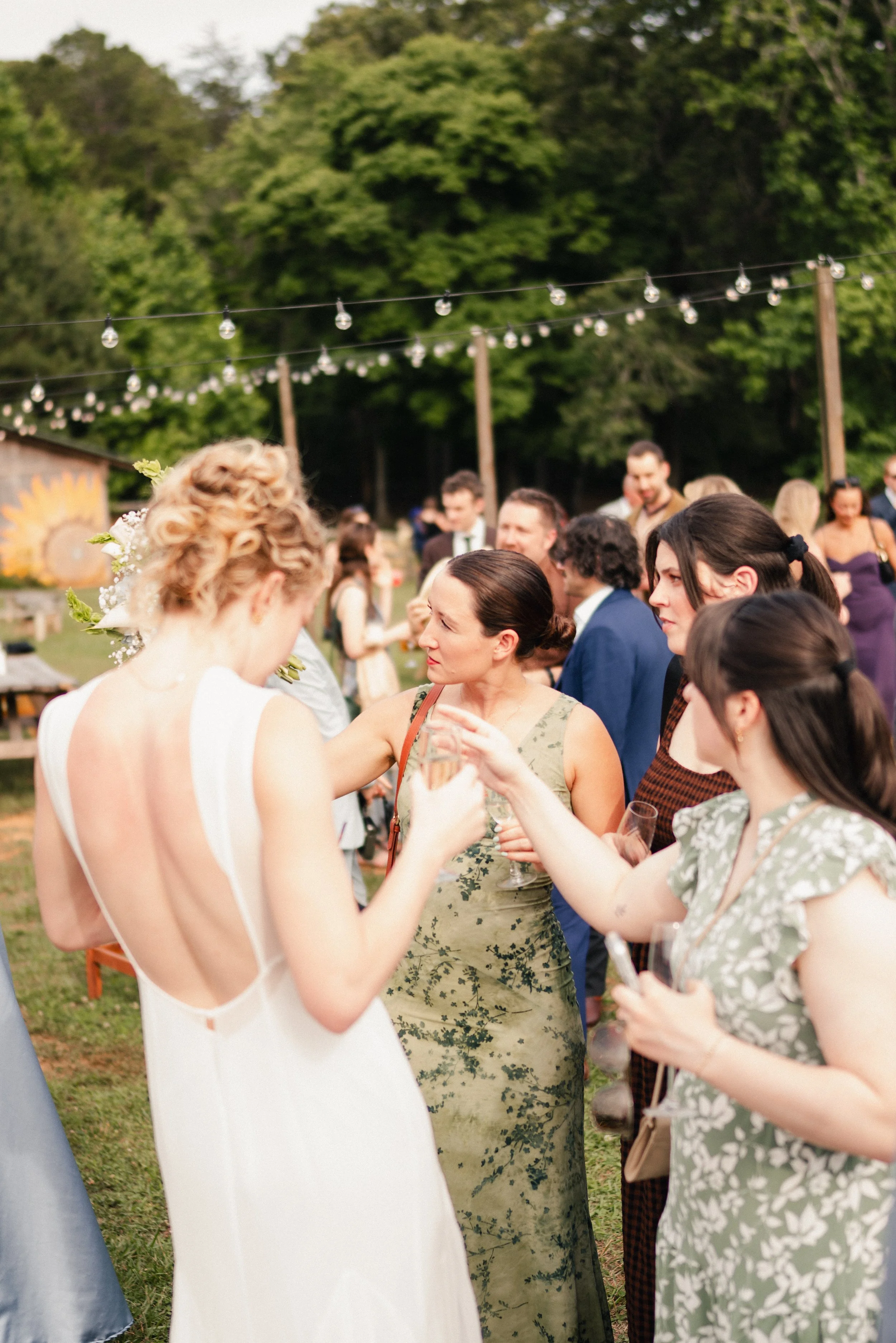 People gathered outdoors at a wedding reception, with some women wearing floral dresses and glasses of champagne, under string lights and surrounded by green trees.
