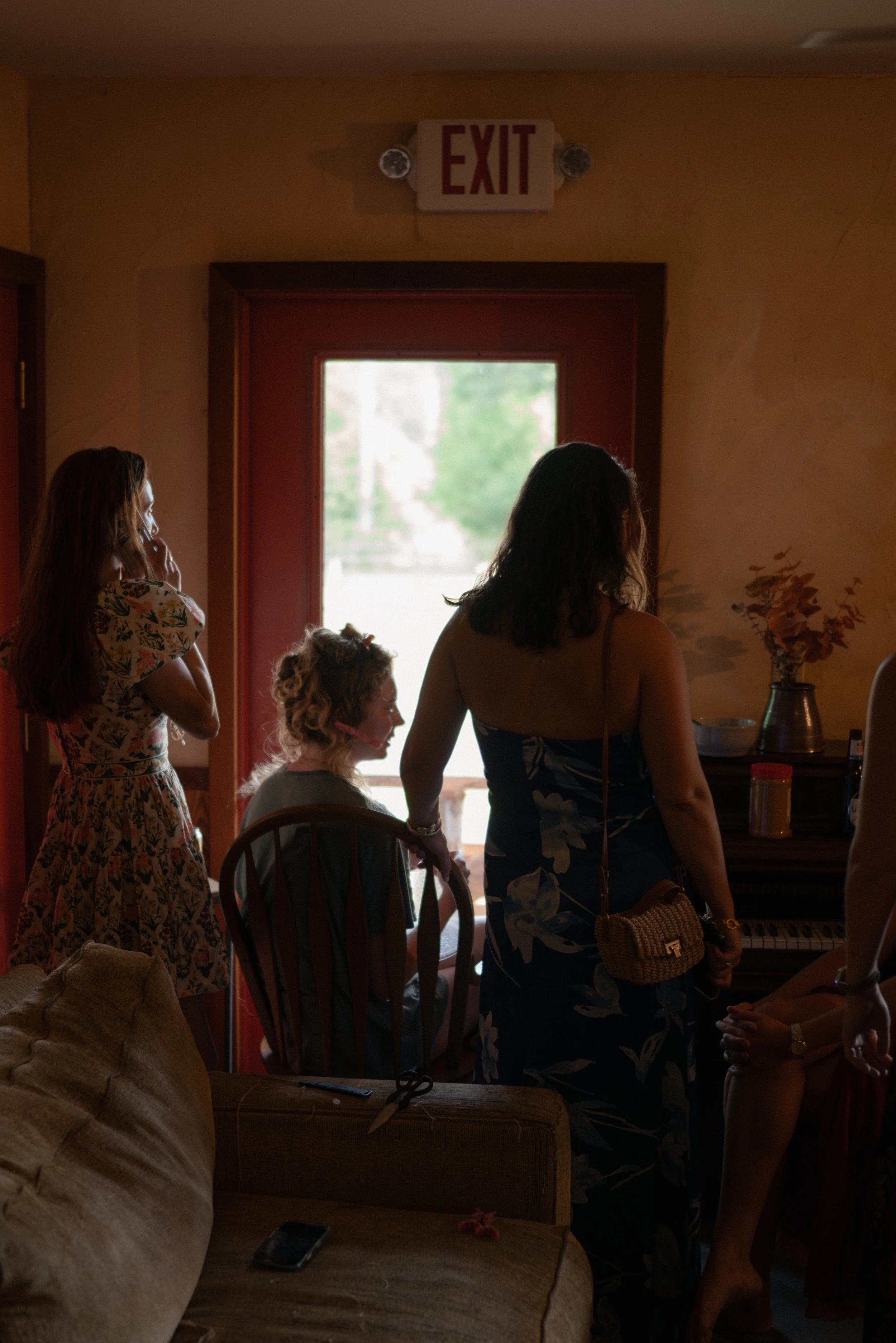 Group of women and a child standing inside near the door, with some sitting on a couch, and a piano visible in the background. Bright outdoor light coming through the door window.