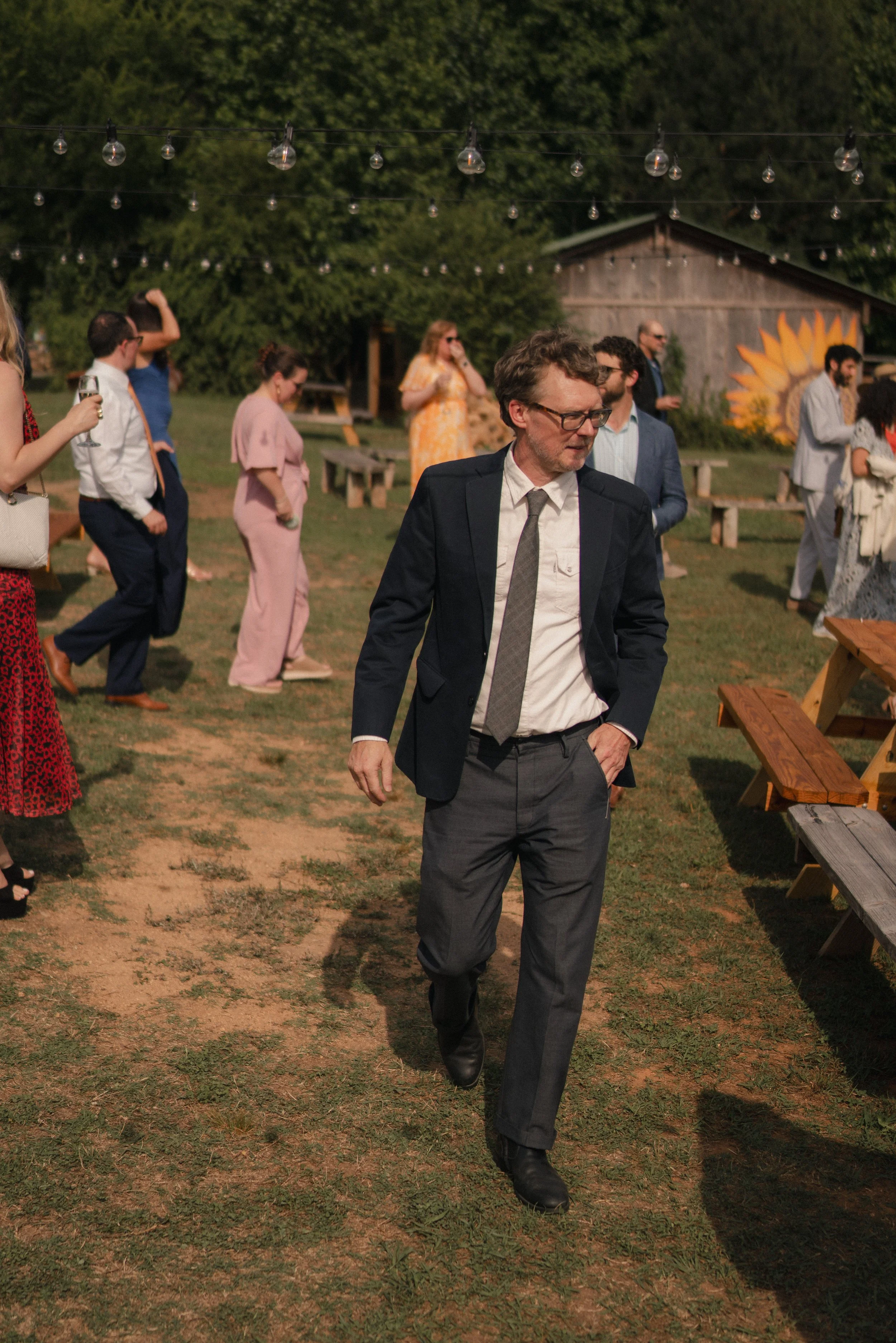 A man in a dark suit and glasses walking through an outdoor gathering with other people socializing under string lights, with a barn and sunflower mural in the background.