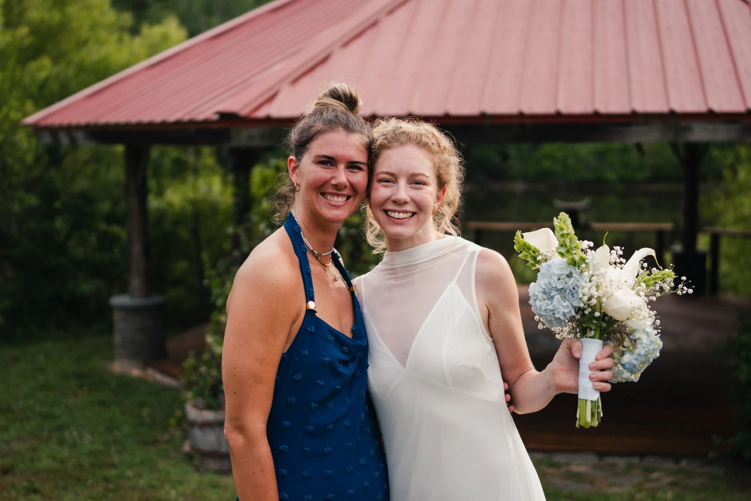 Two women, one in a blue dress and the other in a white wedding dress, smiling and standing outside near a pavilion with a red roof. The woman in white is holding a bouquet of white flowers.