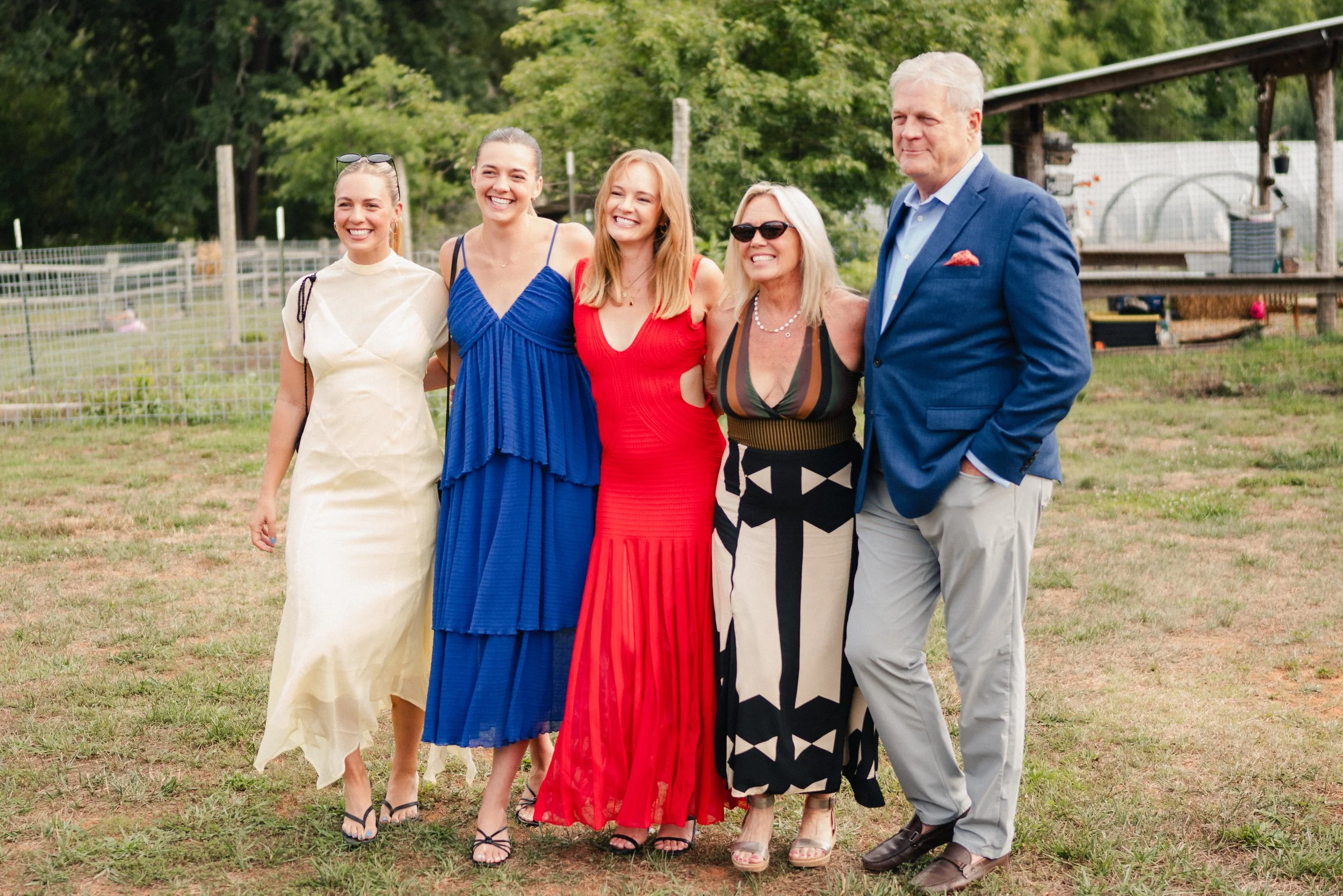 A group of five people, four women and one man, standing outdoors on grass with trees and a barn in the background. They are smiling and dressed in colorful, semi-formal summer clothing.