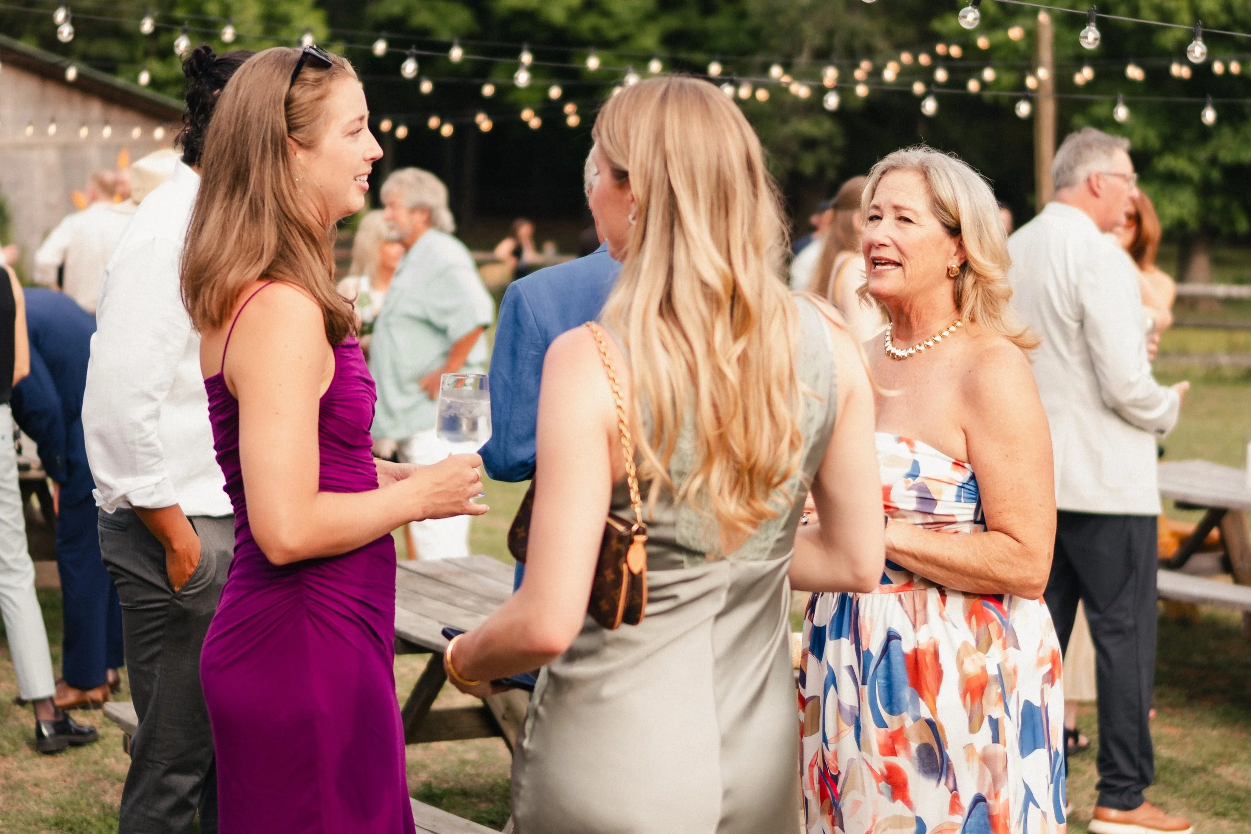 People socializing at an outdoor gathering, with string lights overhead and a wooden fence in the background.
