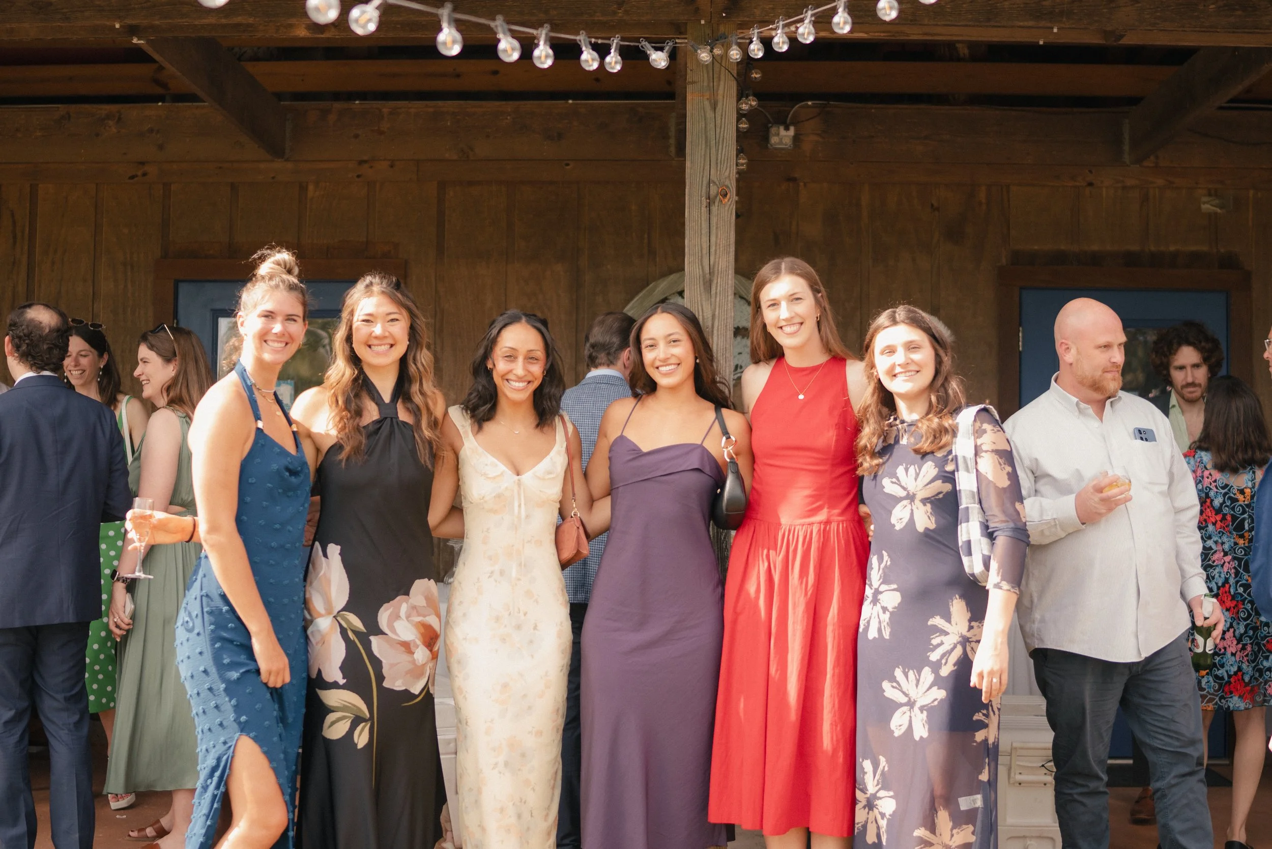 A group of women and men gather at a social event, standing outdoors under string lights, dressed in colorful summer dresses and casual shirts.