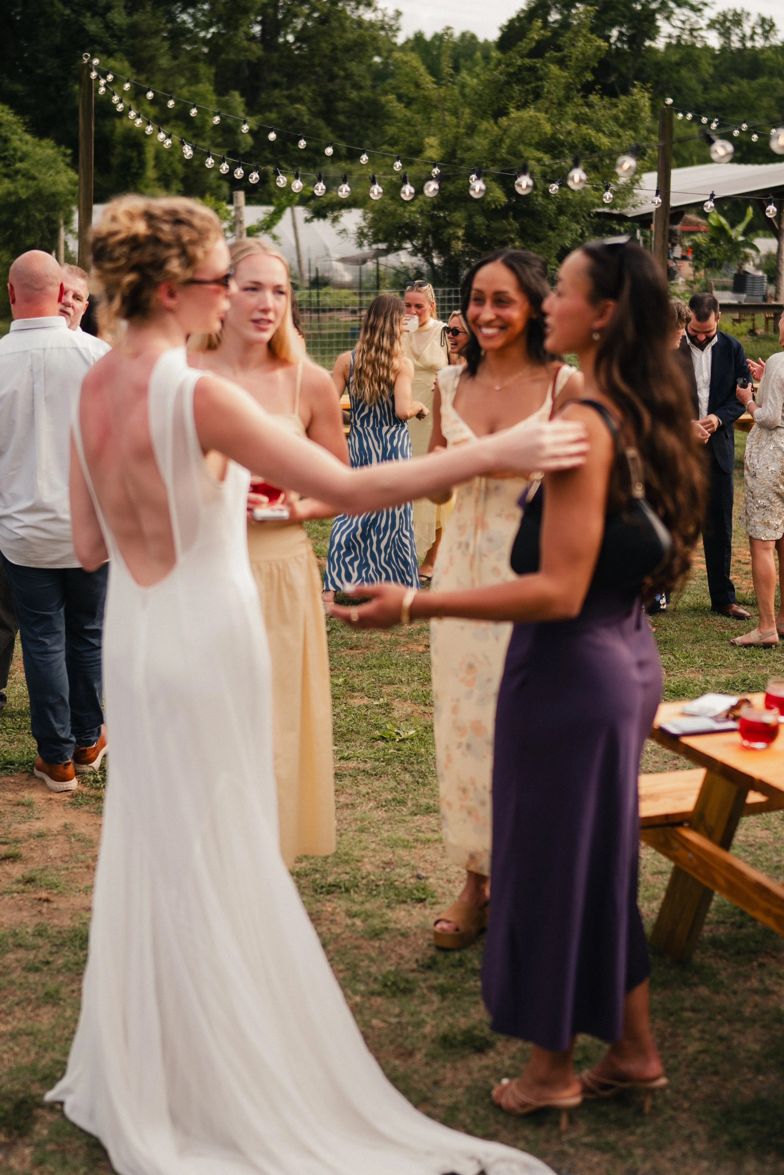 Group of women and men socializing outdoors at sunset, some women wearing dresses, string lights hanging overhead.