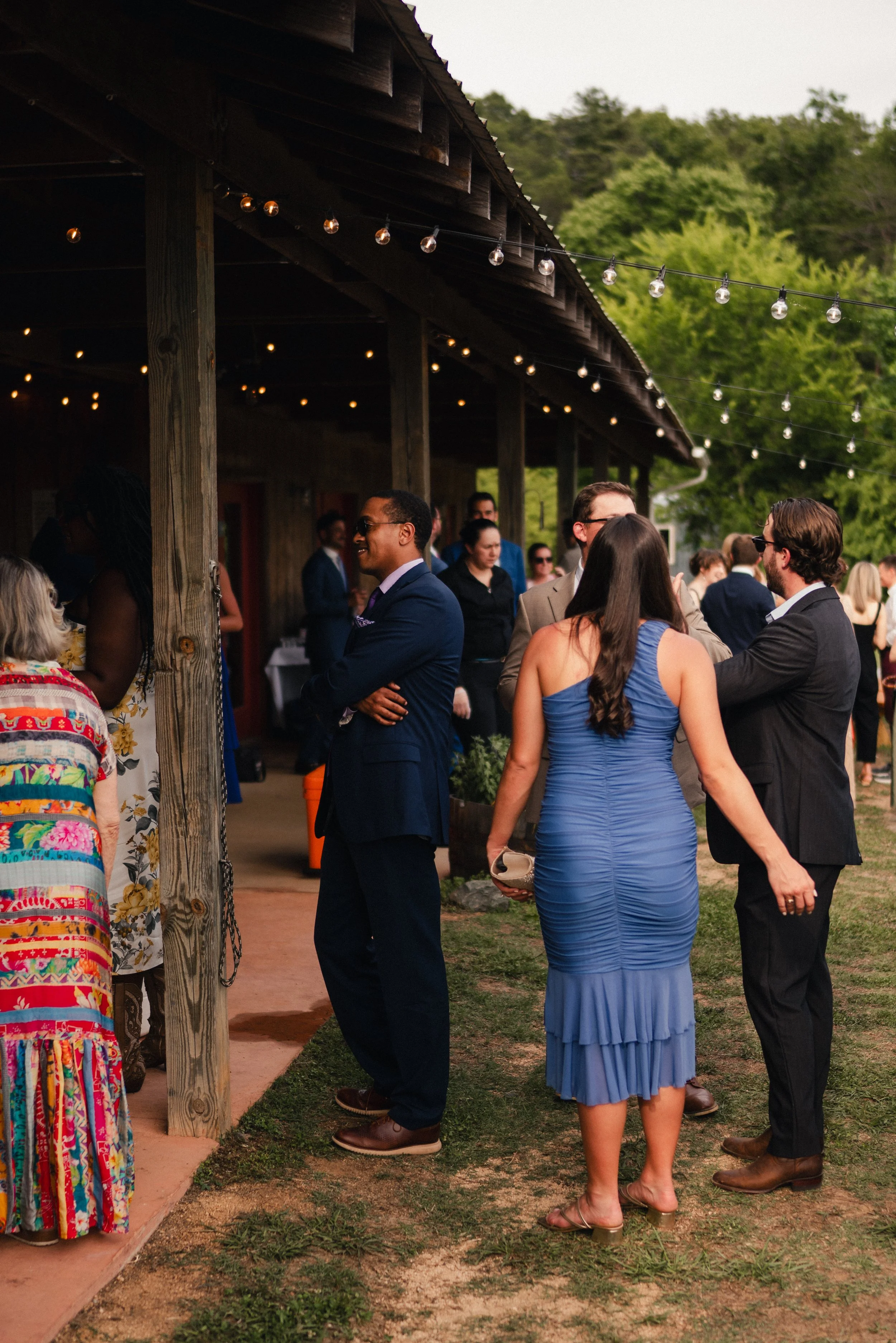 People socializing outdoors during dusk at a gathering with string lights, wooden building, and trees in the background.
