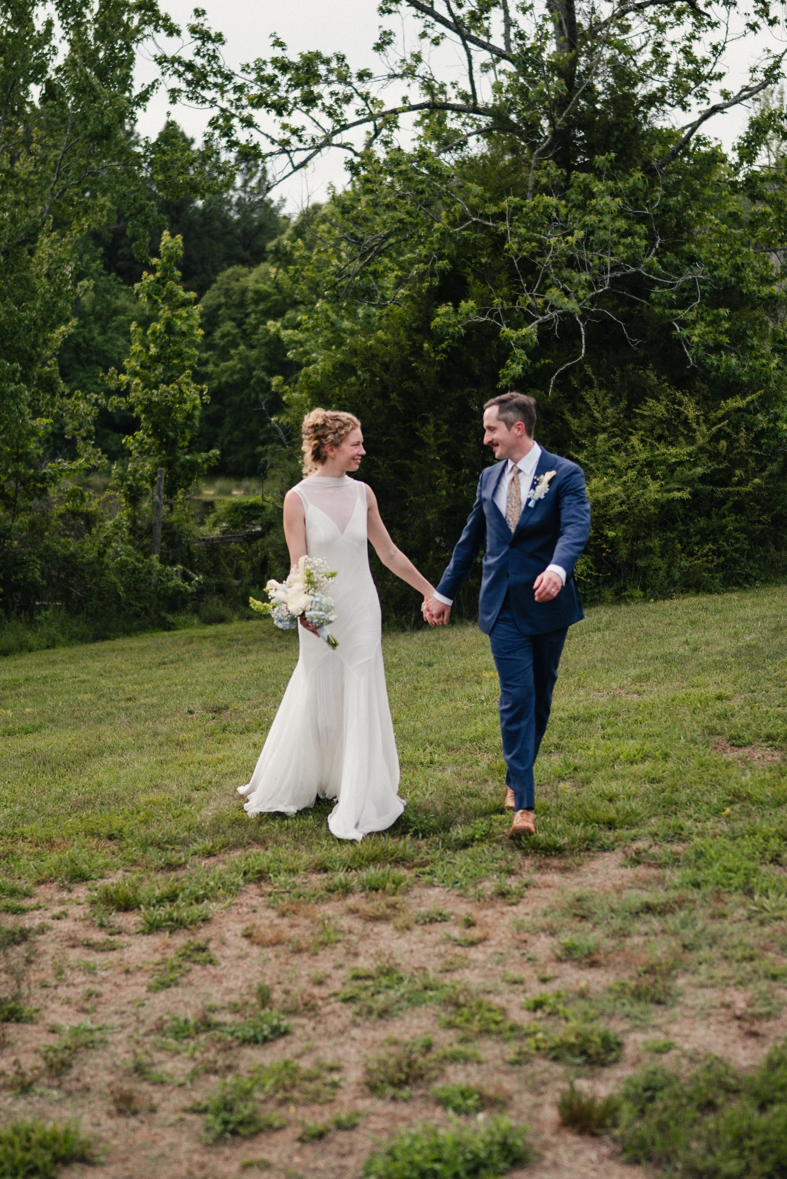 A bride and groom walking hand in hand outdoors on a grassy area, surrounded by green trees, during a wedding ceremony.