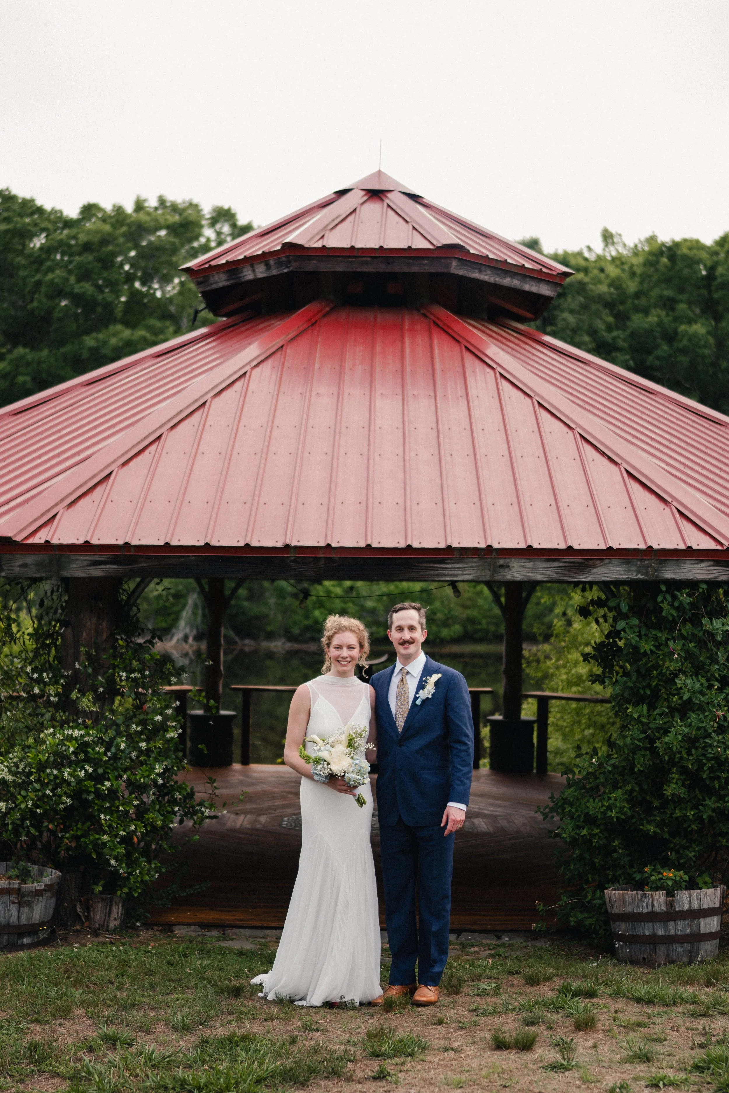 A bride and groom stand together outdoors under a red metal roof pavilion, smiling for a wedding photo. The bride is holding a bouquet, wearing a white dress, and the groom is in a blue suit with a tie and boutonniere. Greenery and trees are visible 