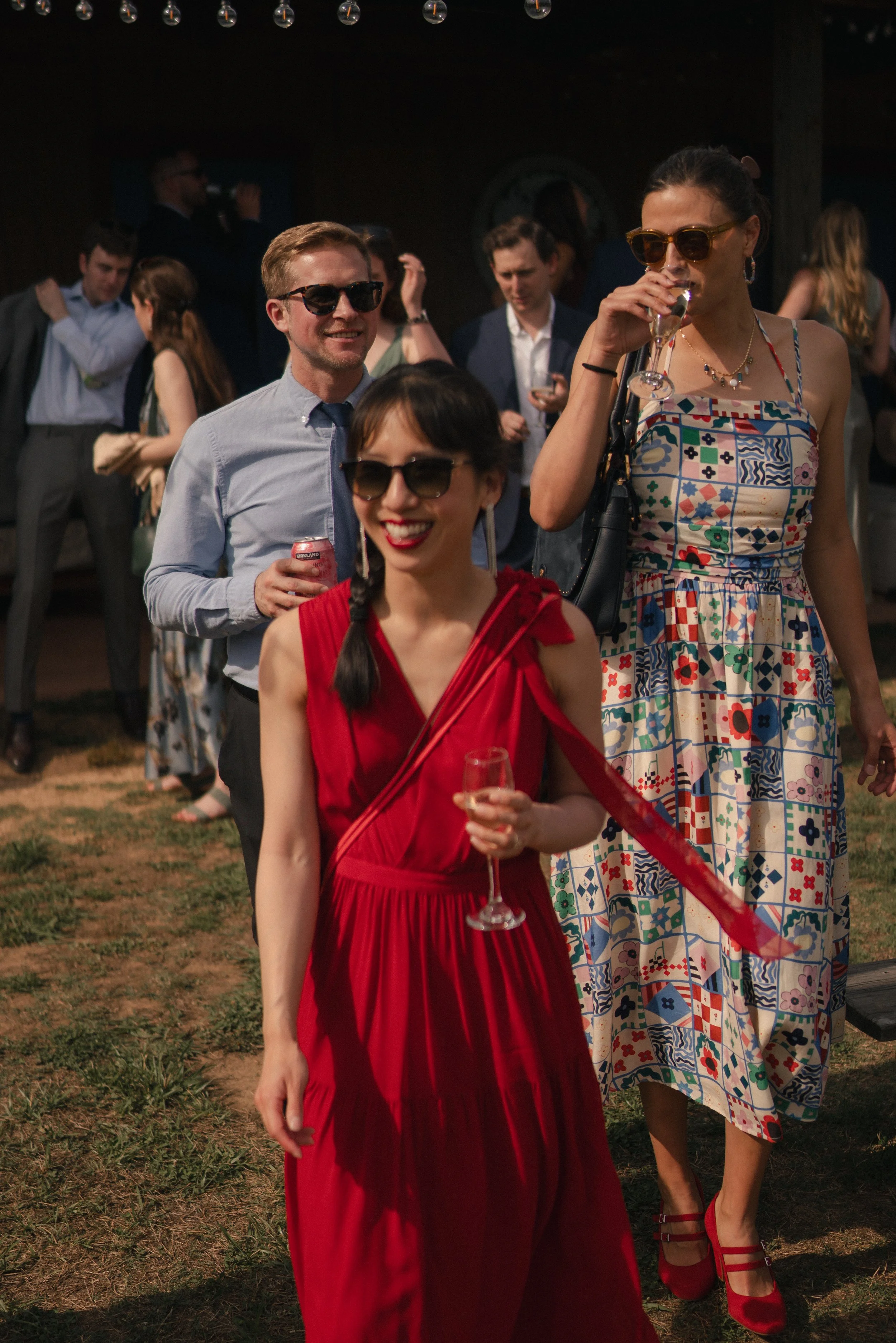 People dressed in vintage clothing at an outdoor party, some wearing sunglasses and holding drinks, with a dark background and string lights overhead.
