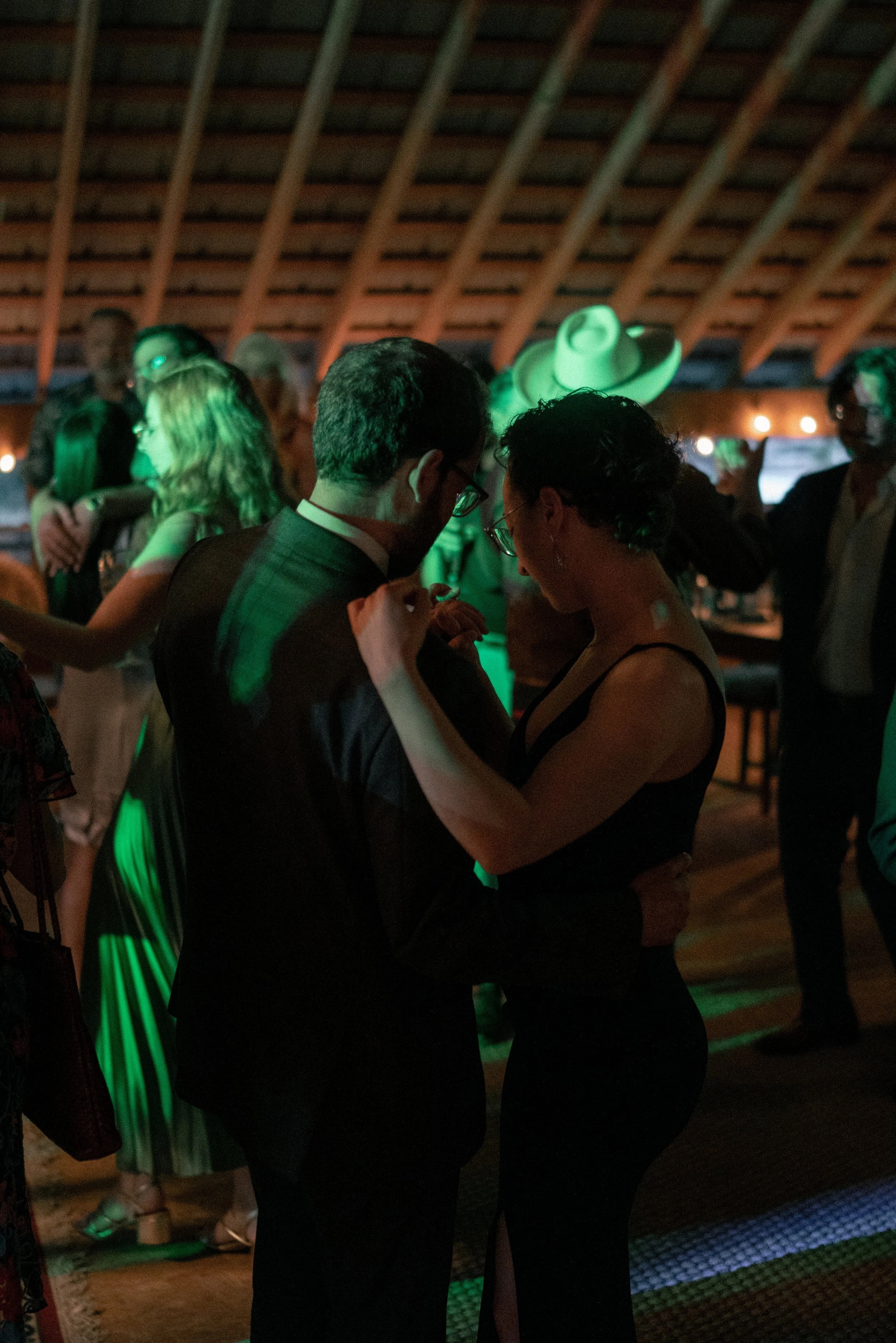 A photo of a couple dancing close together in a dimly lit room with green lighting. The man is wearing a dark suit, and the woman is wearing a black dress with a slit. They are holding hands and leaning into each other.