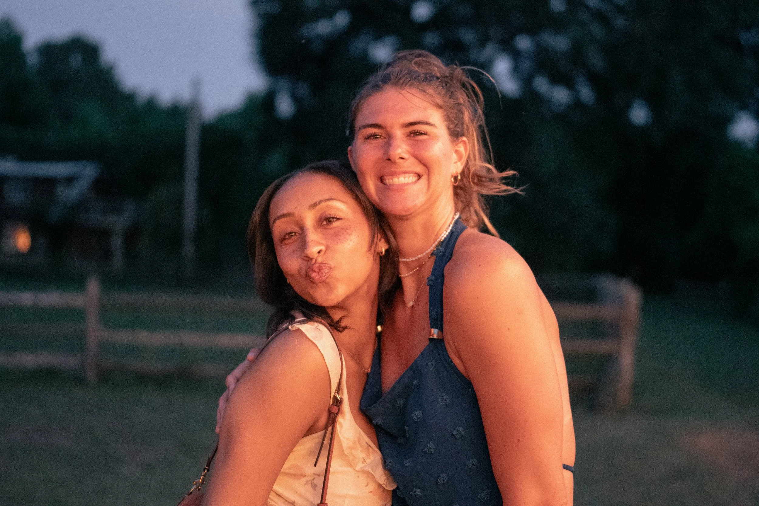 Two young women hugging, smiling, and enjoying time outdoors during sunset with trees and sky in the background.