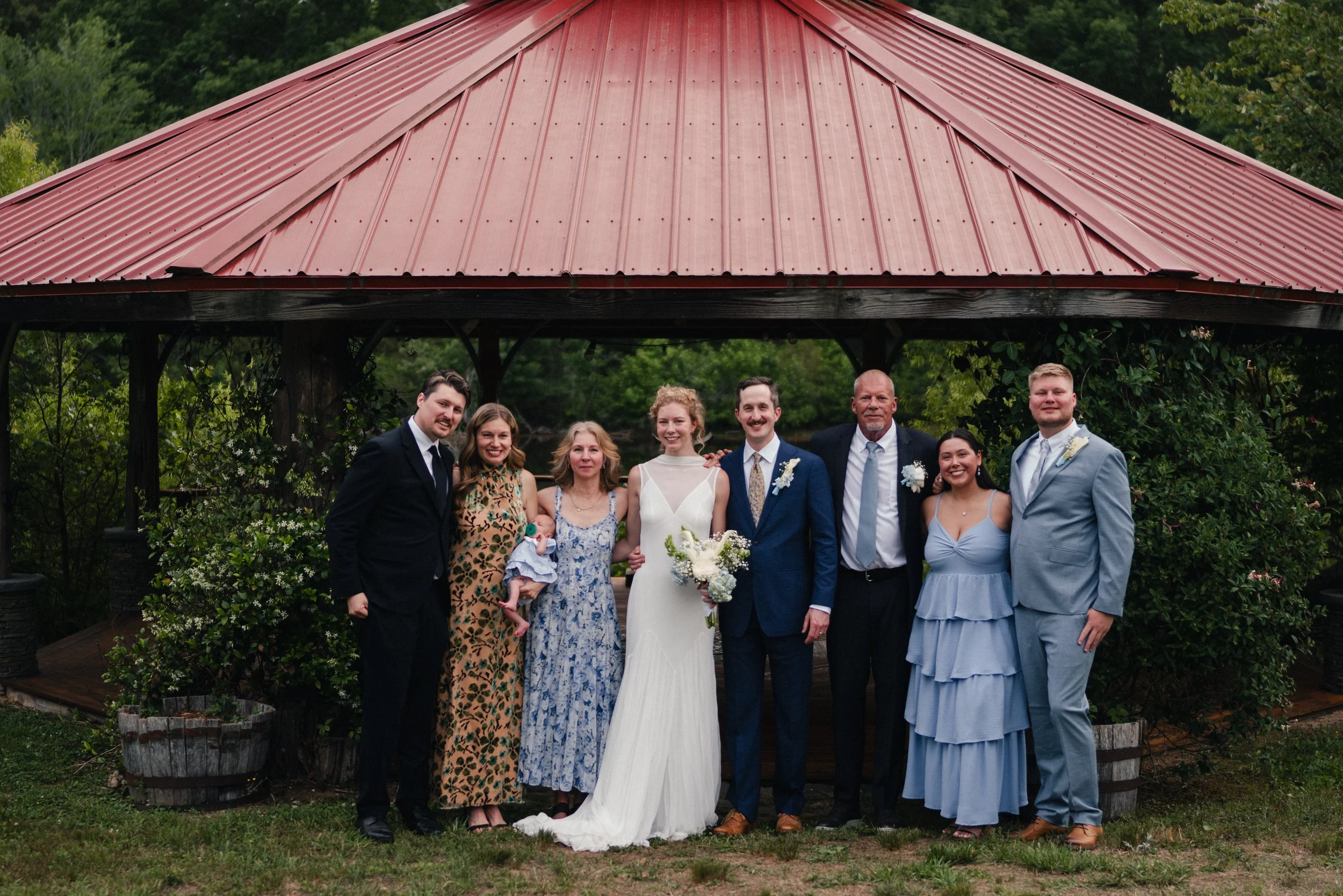Group of people in formal attire standing outdoors under a wooden pavilion with a red metal roof, greenery in background, celebrating a wedding.