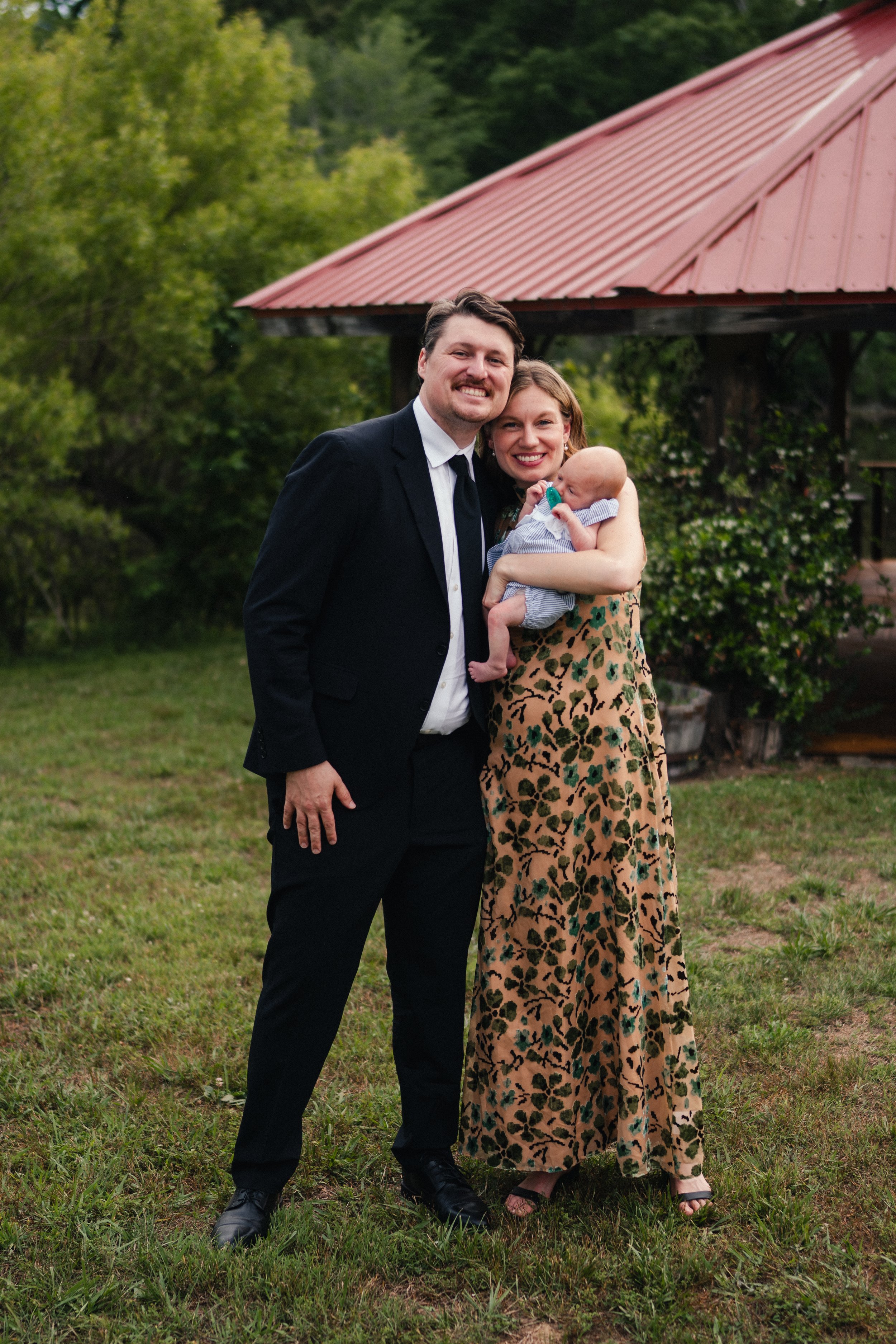 A happy couple holding their baby outdoors in front of a gazebo, with green foliage in the background.