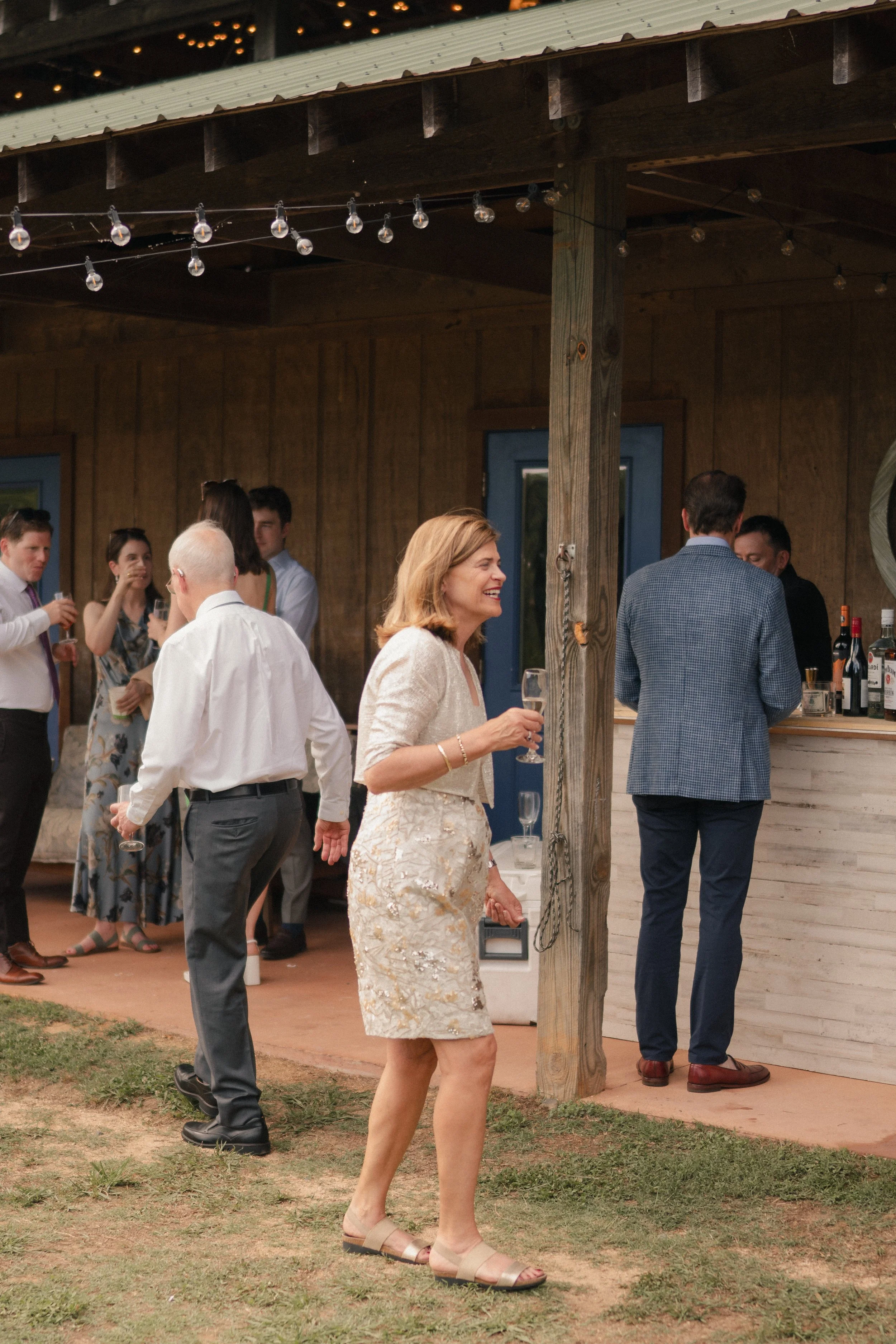 People gathering outdoors at a social event, wine glasses in hand, under a wooden structure with string lights, with a woman in the foreground smiling and wearing a beige dress, and others in the background in casual and semi-formal attire.