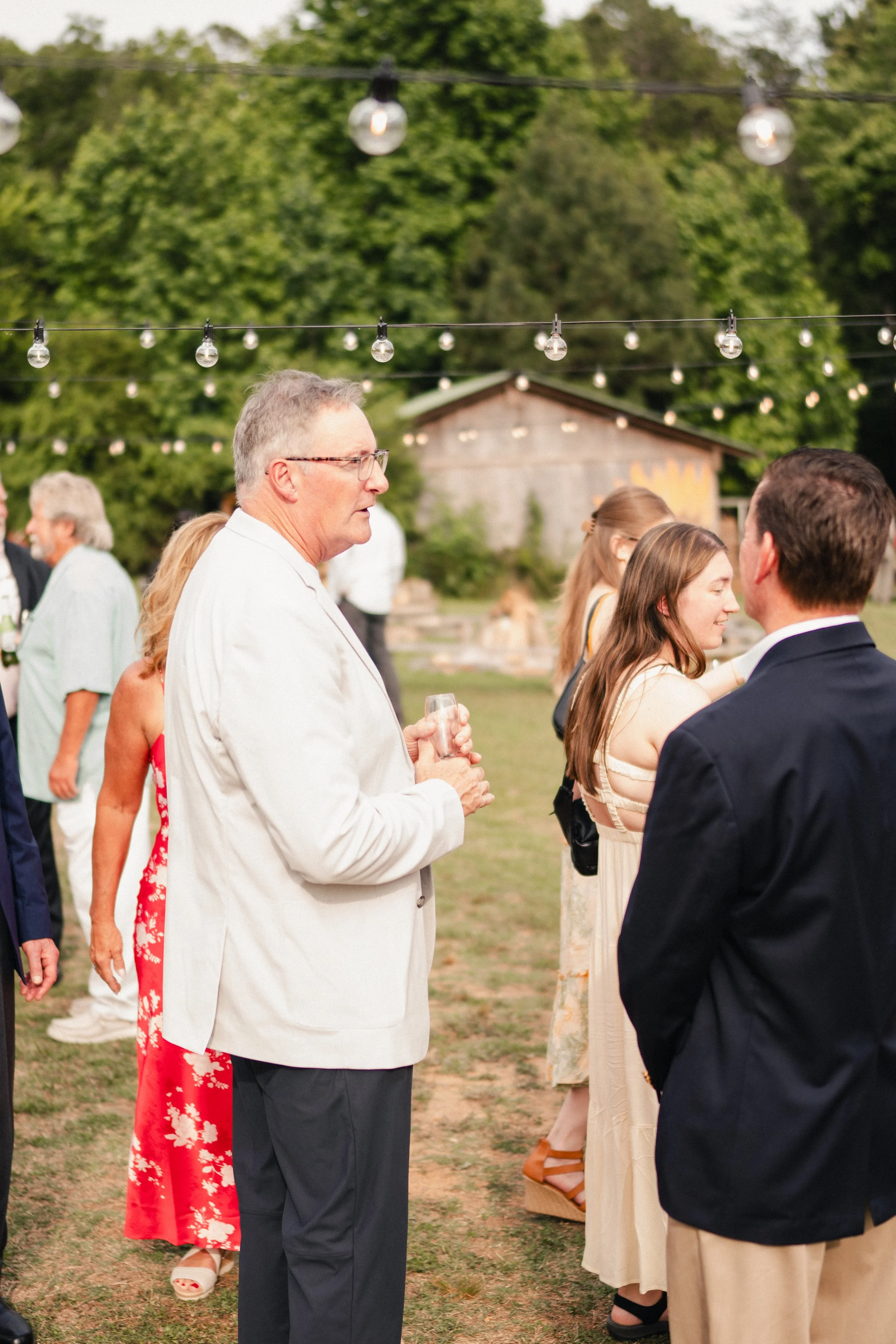 A group of people dressed in semi-formal attire gathered outdoors for a social event or celebration, with string lights hanging overhead and a rustic building and lush green trees in the background.