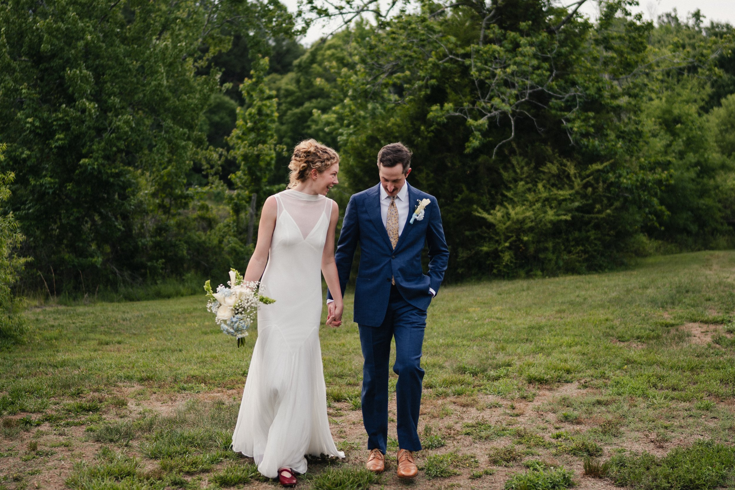A couple, dressed in wedding attire, walks hand in hand on grass with green trees in the background. The bride wears a white gown and holds a bouquet, while the groom is dressed in a blue suit with a boutonniere.