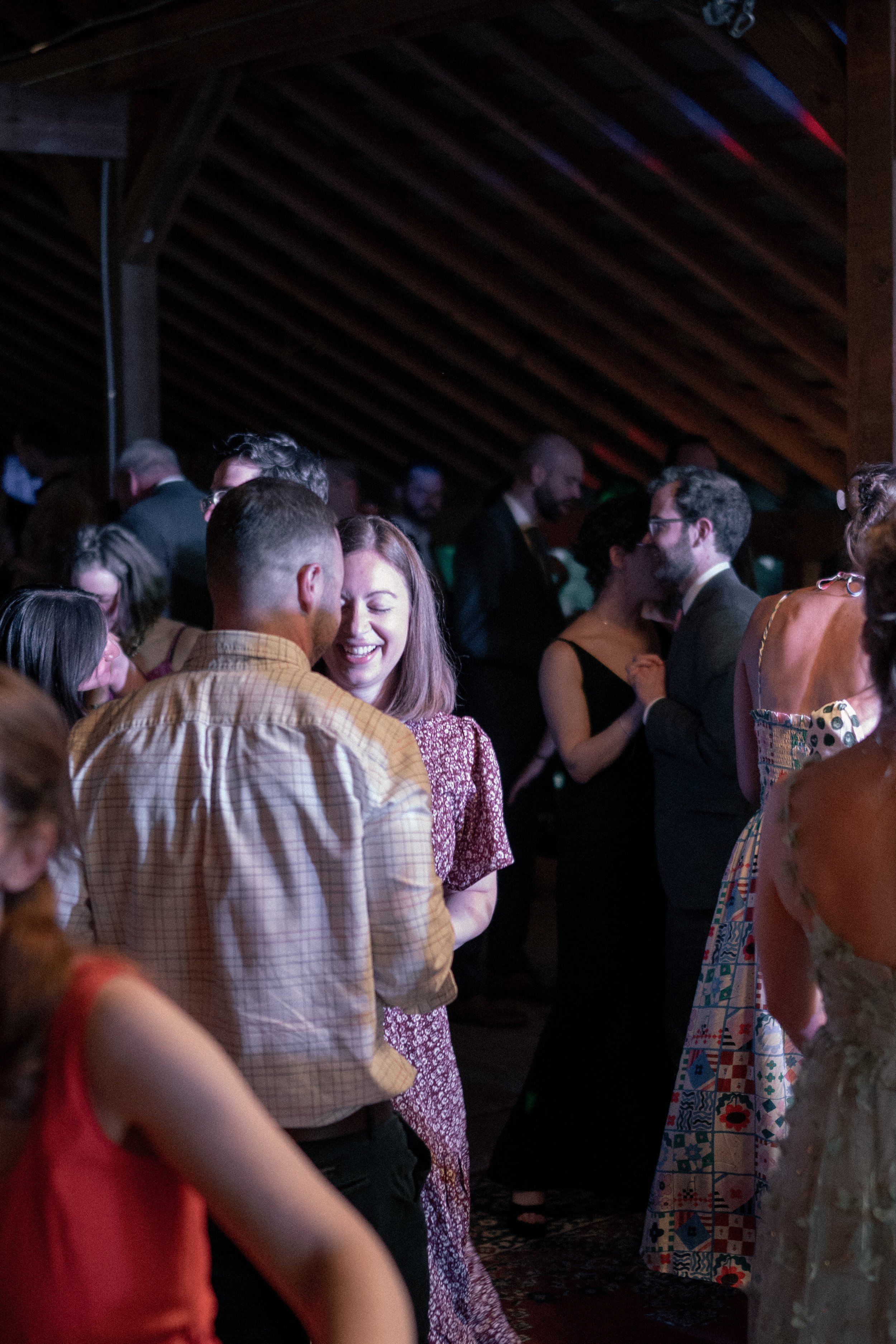 People dancing at a lively social event in a dimly lit room with wooden ceiling beams, wearing colorful and formal attire.