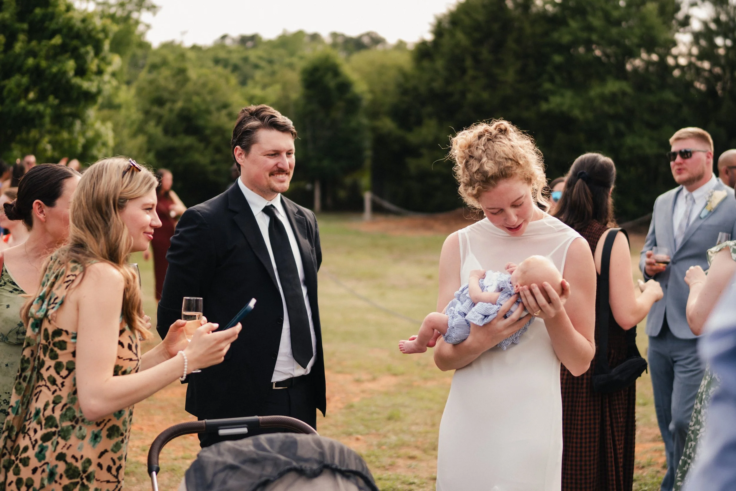A woman in a white dress holding a newborn baby at an outdoor gathering. A man in a suit stands next to her, smiling, with several people in the background. The event appears to be a celebration or ceremony, set in a green, lush outdoor area.