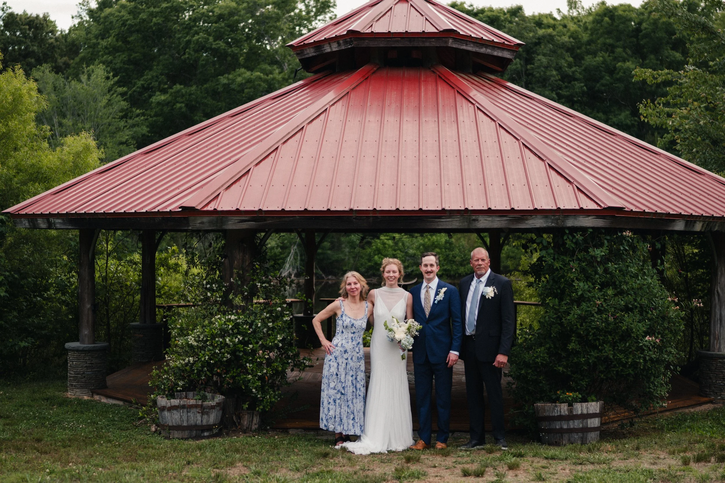 Group of four people, three adults and one bride, standing under a red metal-roofed gazebo surrounded by lush green trees. The bride is holding a bouquet and wearing a white wedding dress. The others are dressed in formal attire, with the women in dr