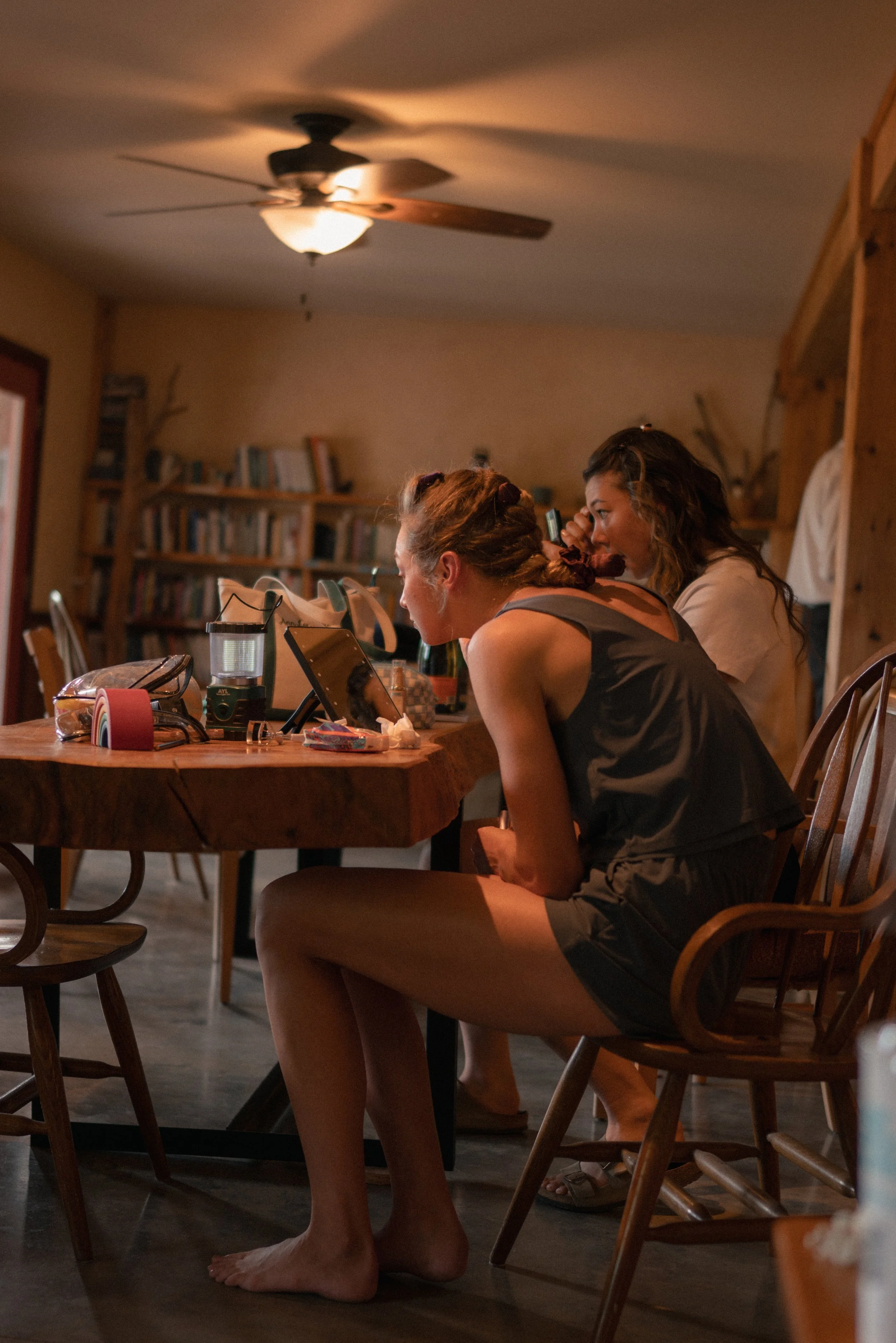 Two women sitting at a wooden table, engaged in a conversation, with one woman in a gray top and a woman in a white shirt. The table has various items including a tablet, makeup, and personal belongings. The room has a warm, cozy atmosphere with wood