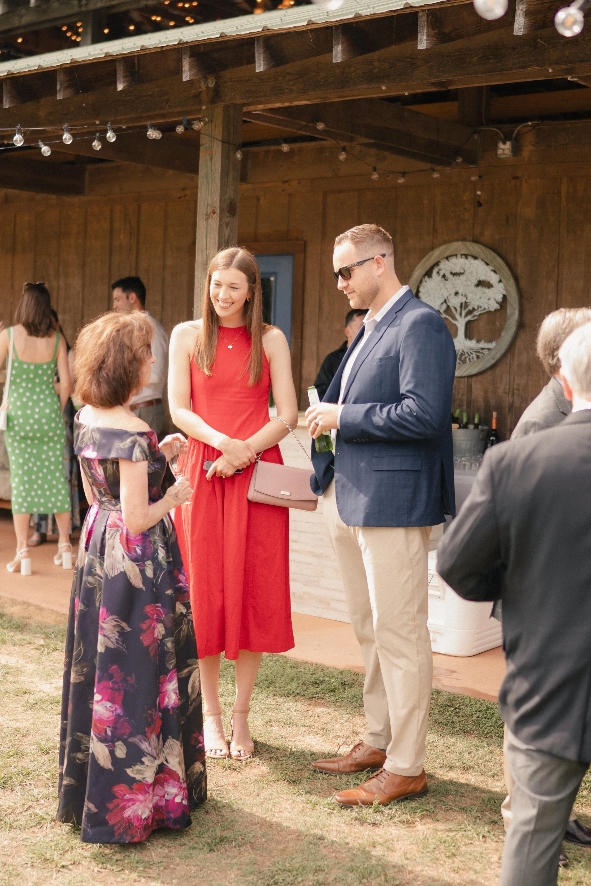 People gathered outdoors at an event, engaged in conversation, with a wooden backdrop, string lights, and a decorative tree artwork on the wall.