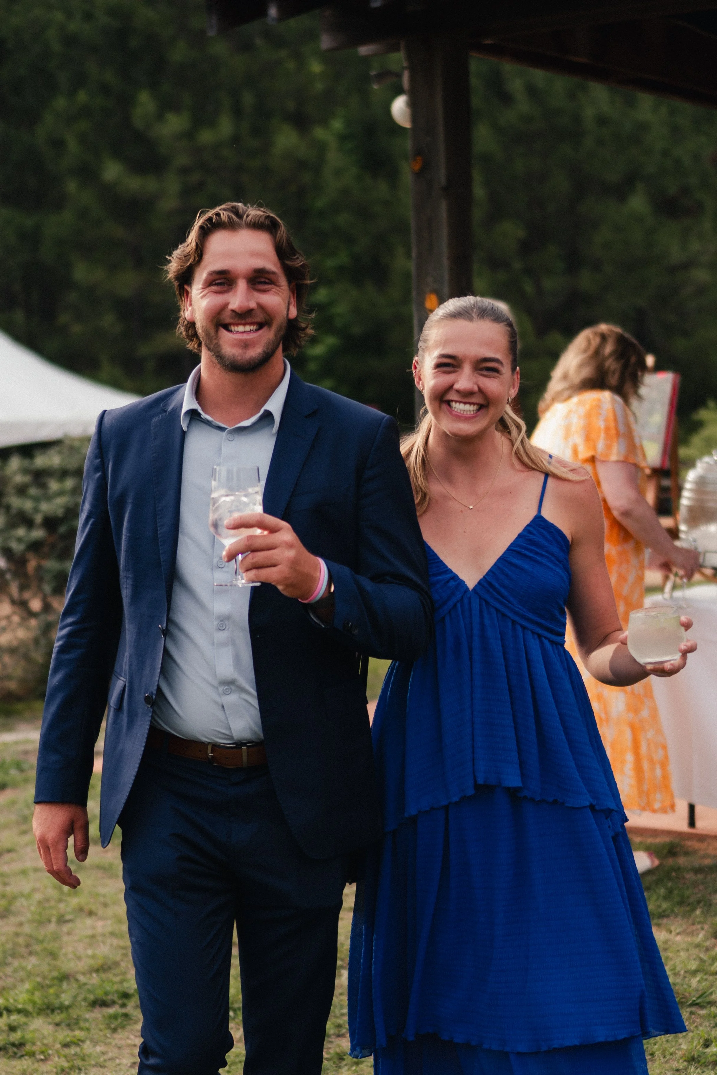 A smiling man in a navy suit and a woman in a blue dress enjoying a social gathering outdoors with trees and a tent in the background.