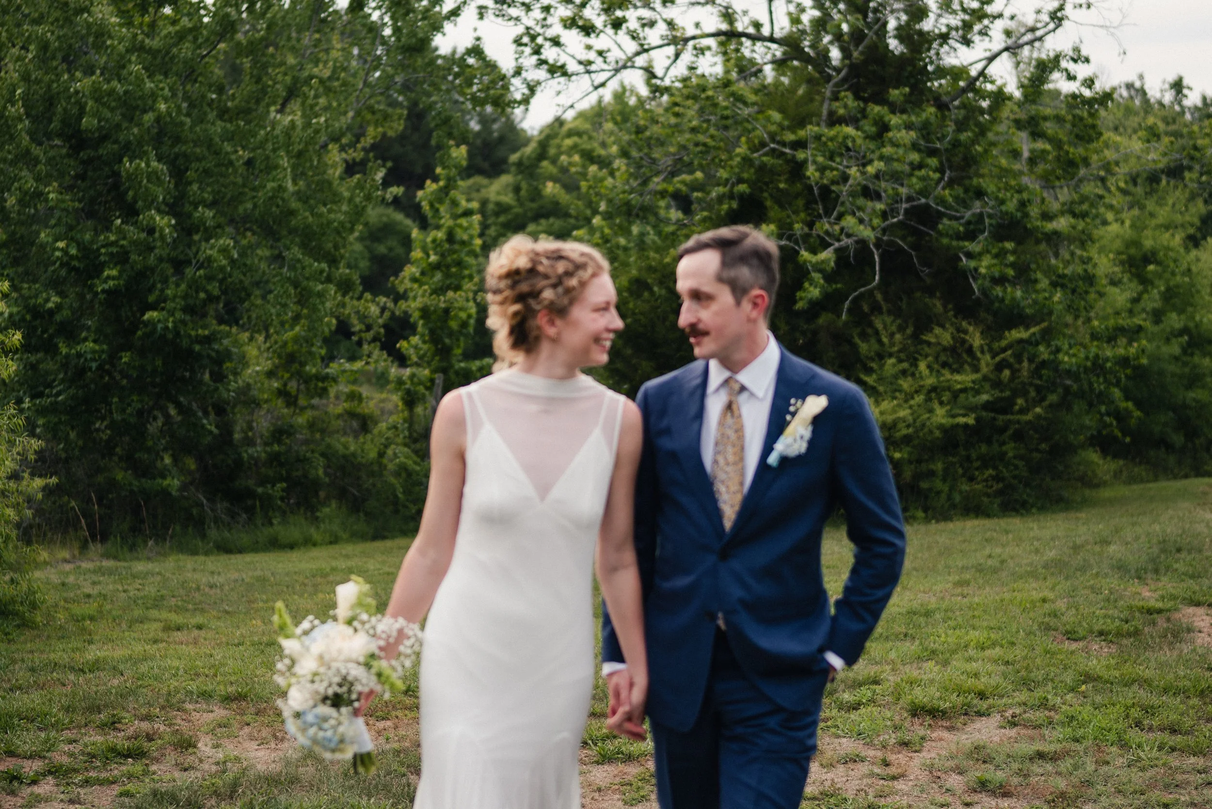 A bride and groom walking outdoors on grass, holding hands and smiling at each other, with trees and greenery in the background.