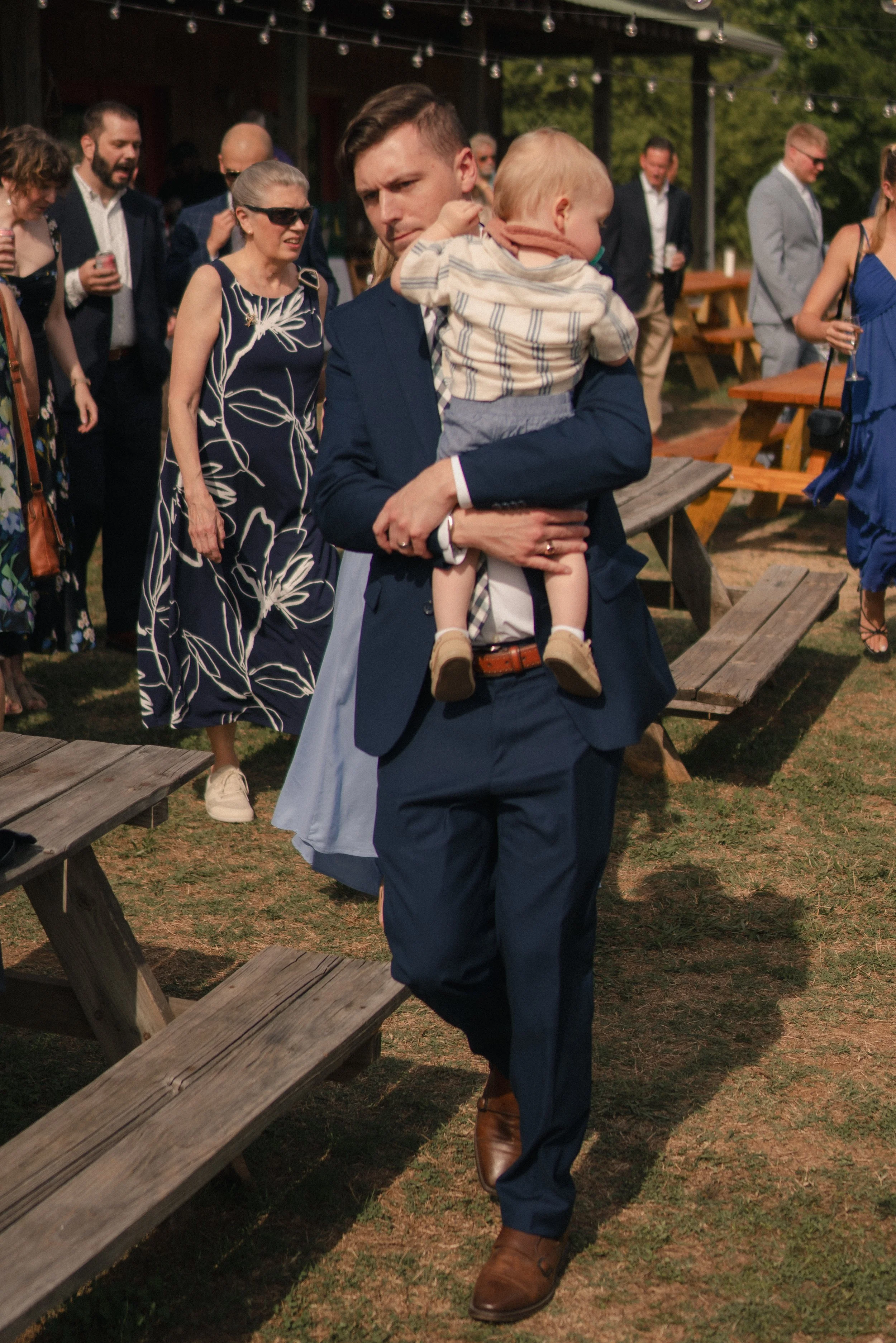 A man in a navy suit and brown shoes is holding a young child dressed in a striped shirt and shorts. The scene is outdoors with several people dressed in semi-formal clothing, some holding drinks, and wooden picnic tables in the background. The atmos