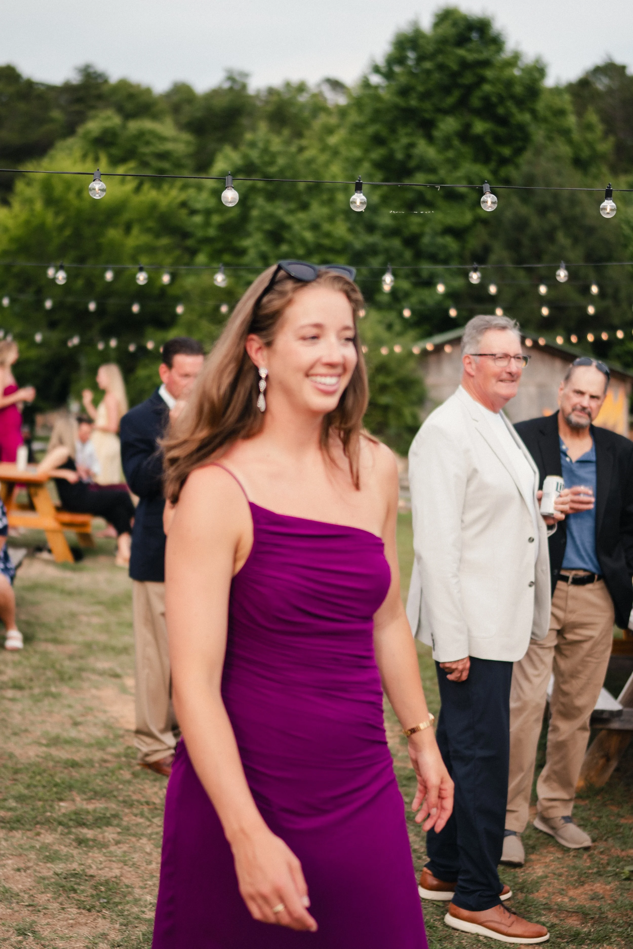A woman in a purple dress smiling at an outdoor gathering, with string lights overhead and other people in the background