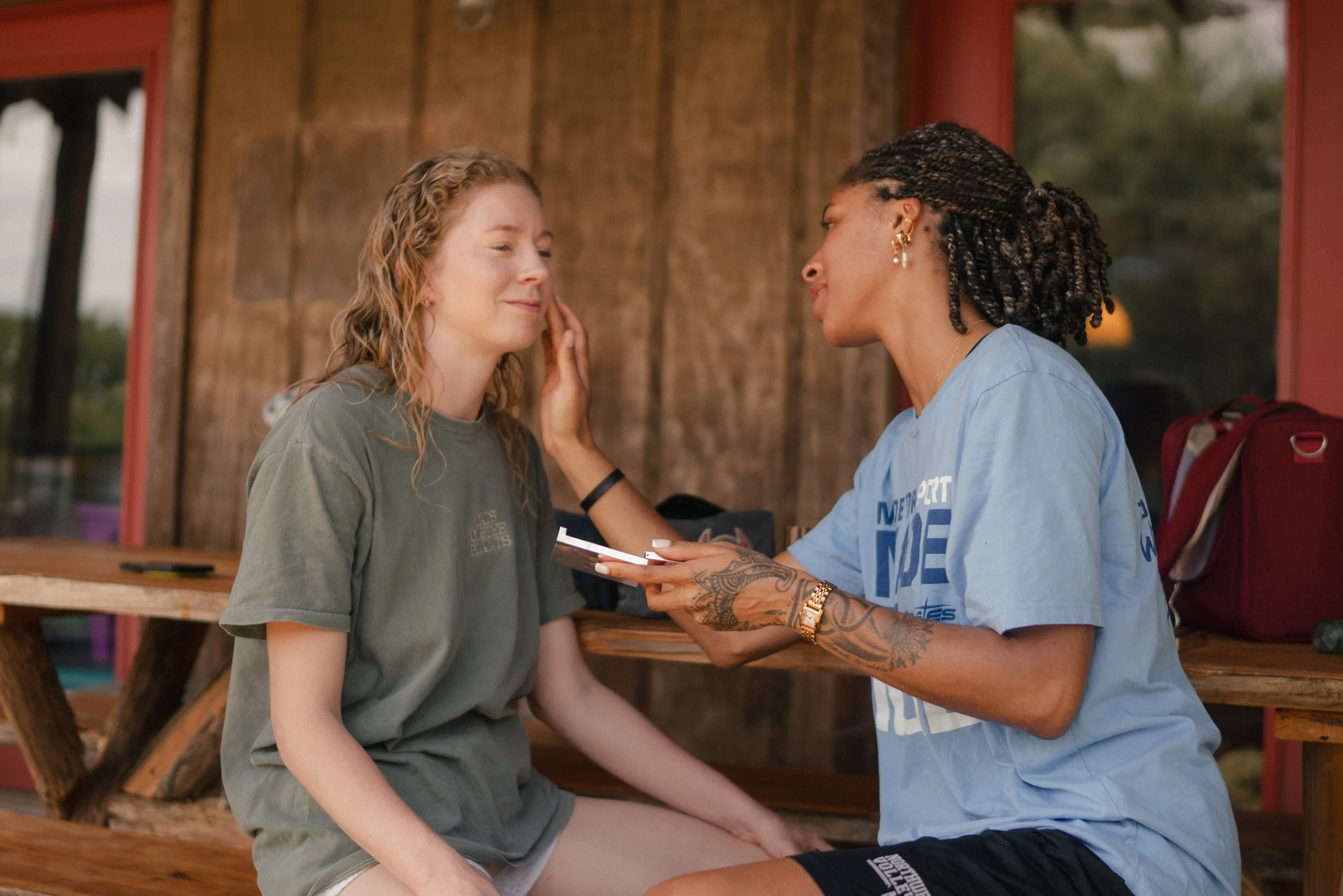 Two women with contrasting hairstyles and skin tones sitting and looking at each other, with one woman touching the other's face.