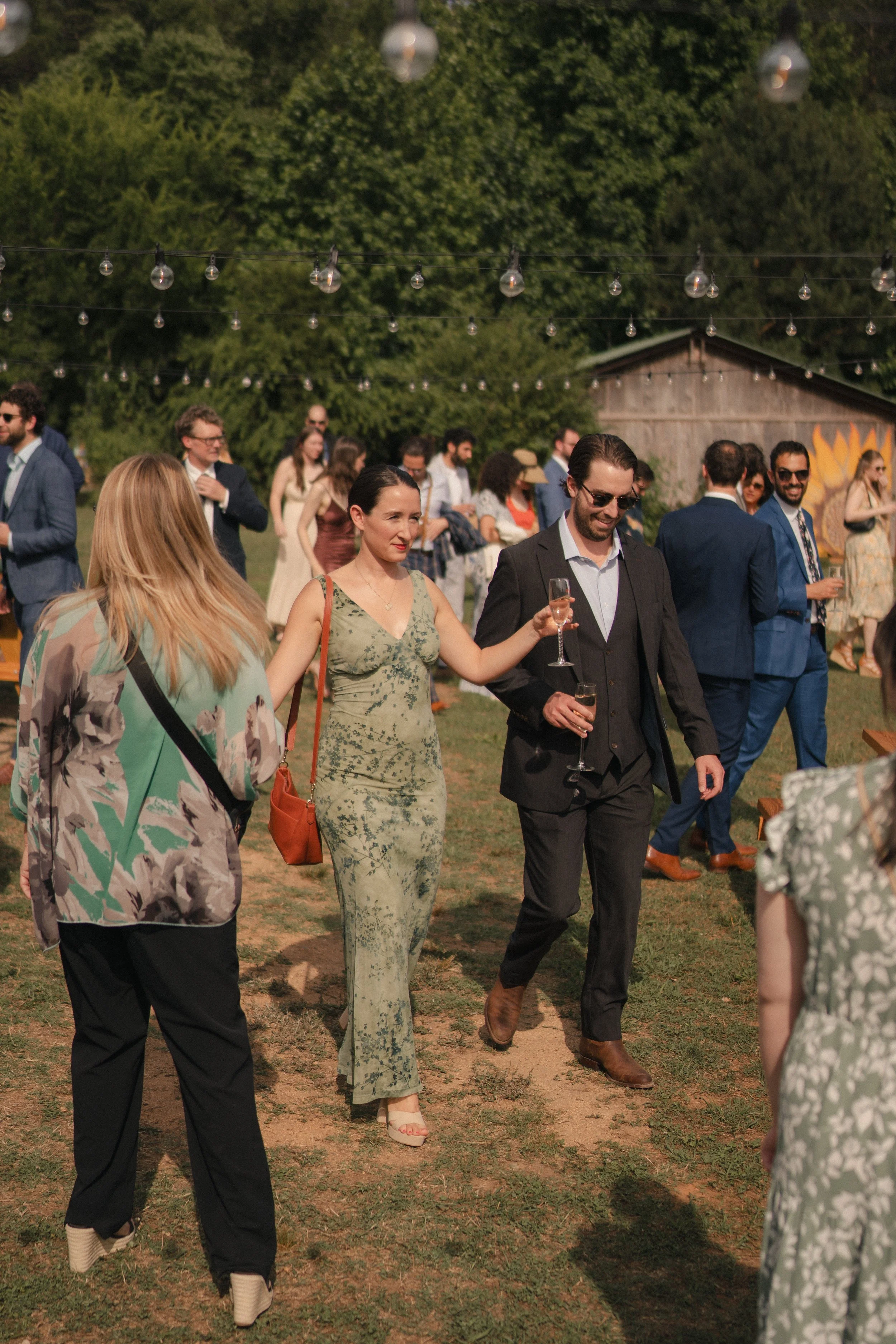 A group of people dressed in formal attire at an outdoor event, standing on grass under string lights, with trees and a shed in the background.