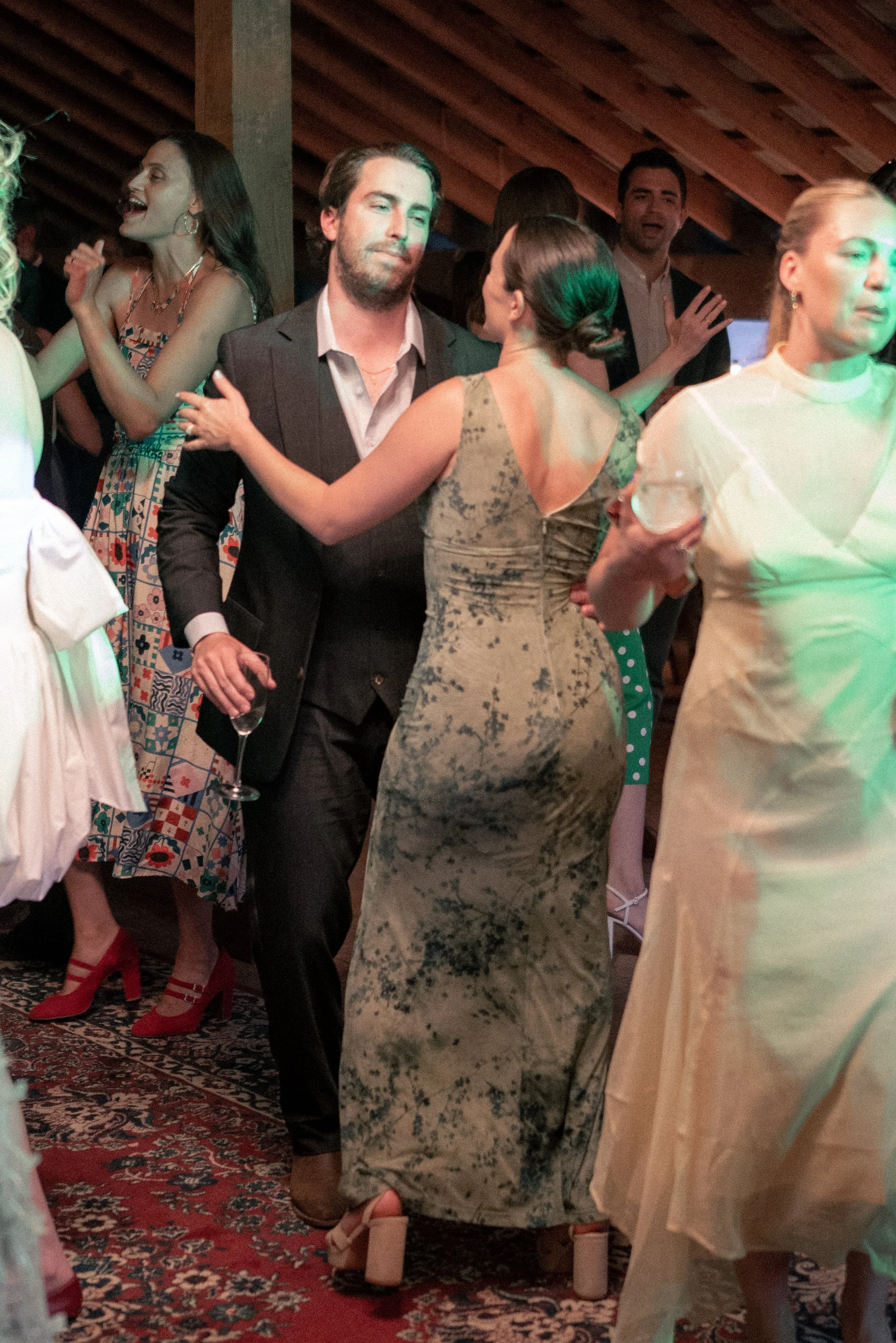 People dancing at a party on a patterned carpet, with a wooden ceiling overhead. A woman in a patterned dress and red shoes dances with a man in a black suit. A woman in a green dress and heels holds a drink nearby, and others are seen enjoying the e