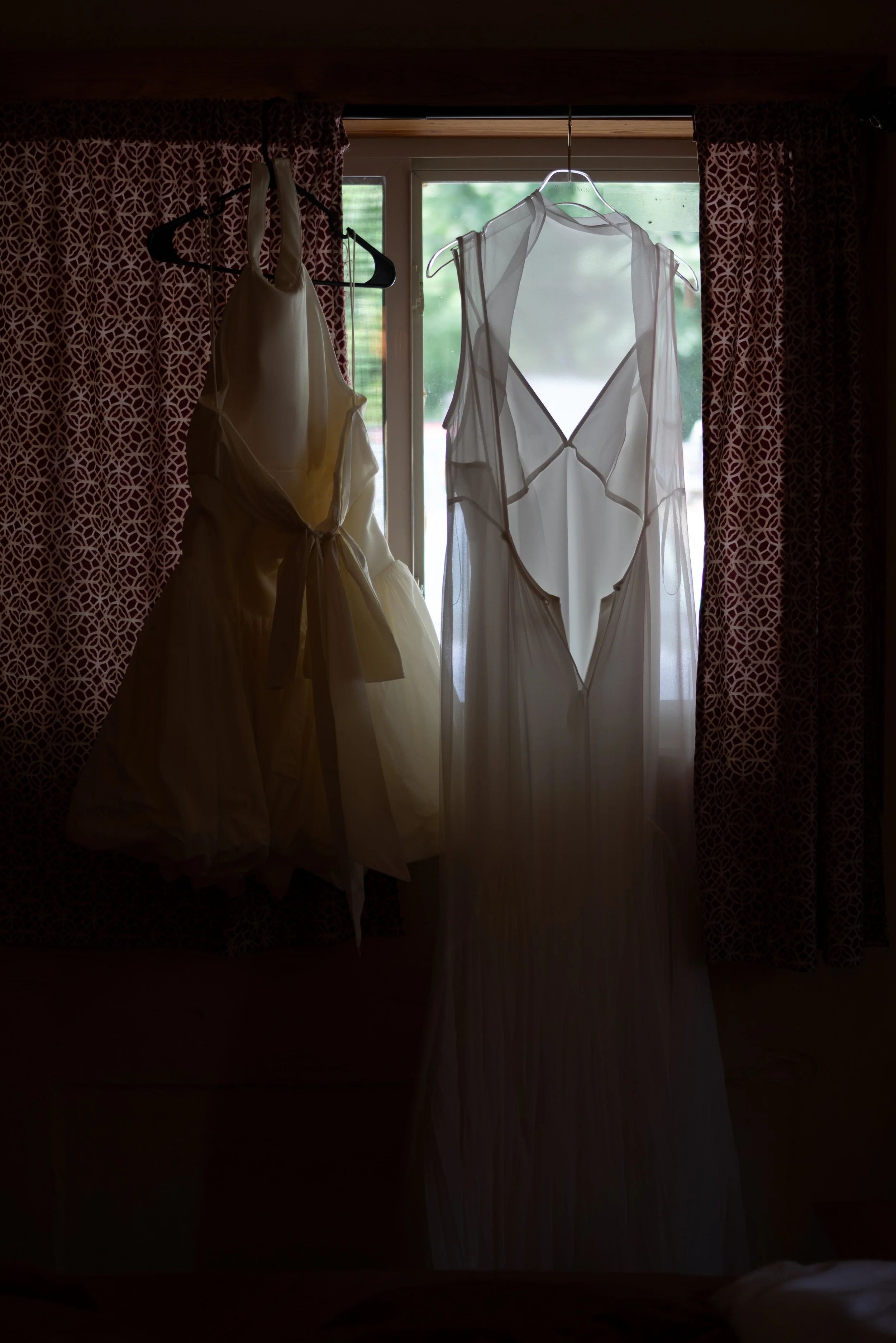 Two dresses hanging on hangers in front of a window with curtains on either side.