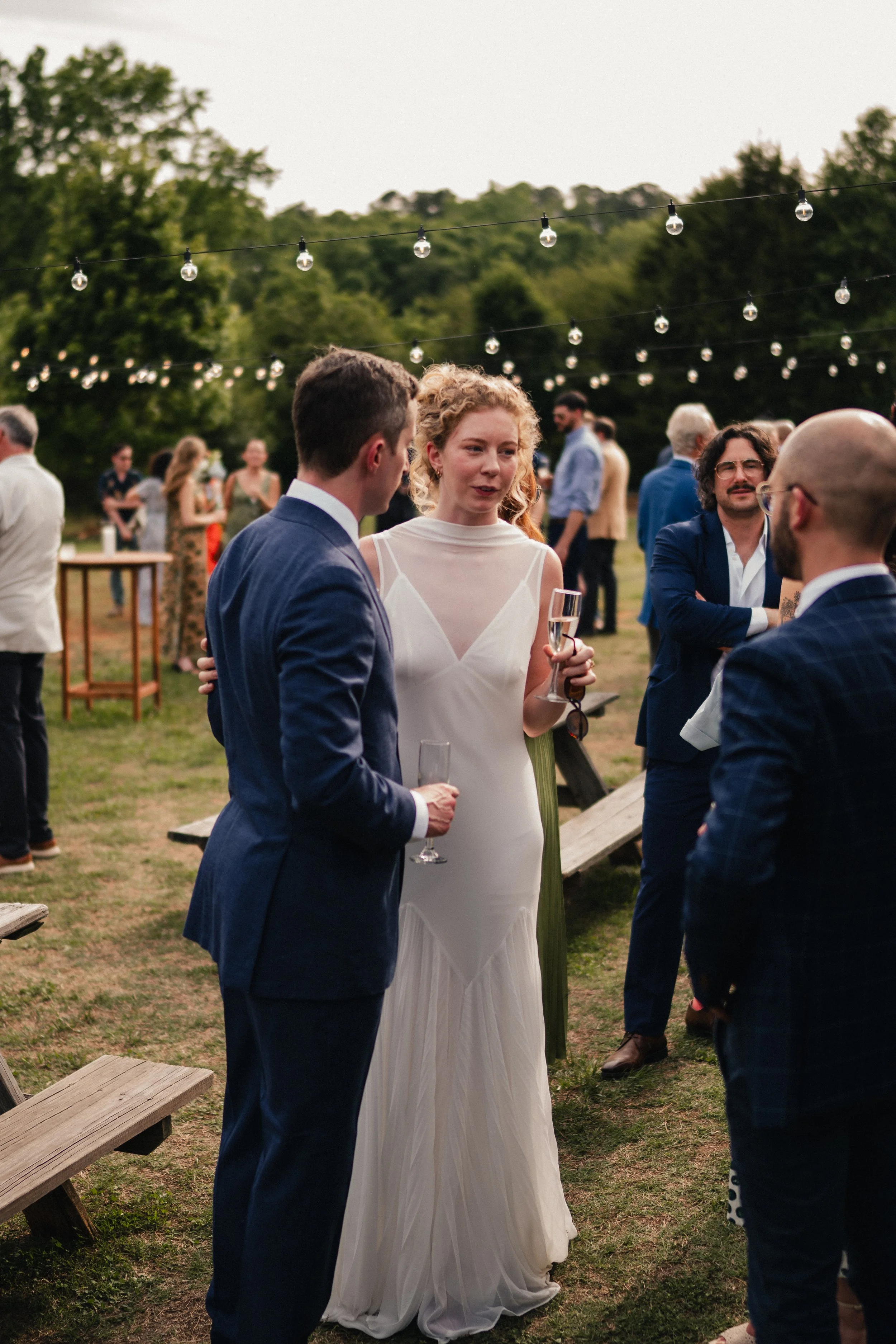 Outdoor wedding reception with string lights, guests in formal attire, and a woman in a white dress holding a champagne flute.
