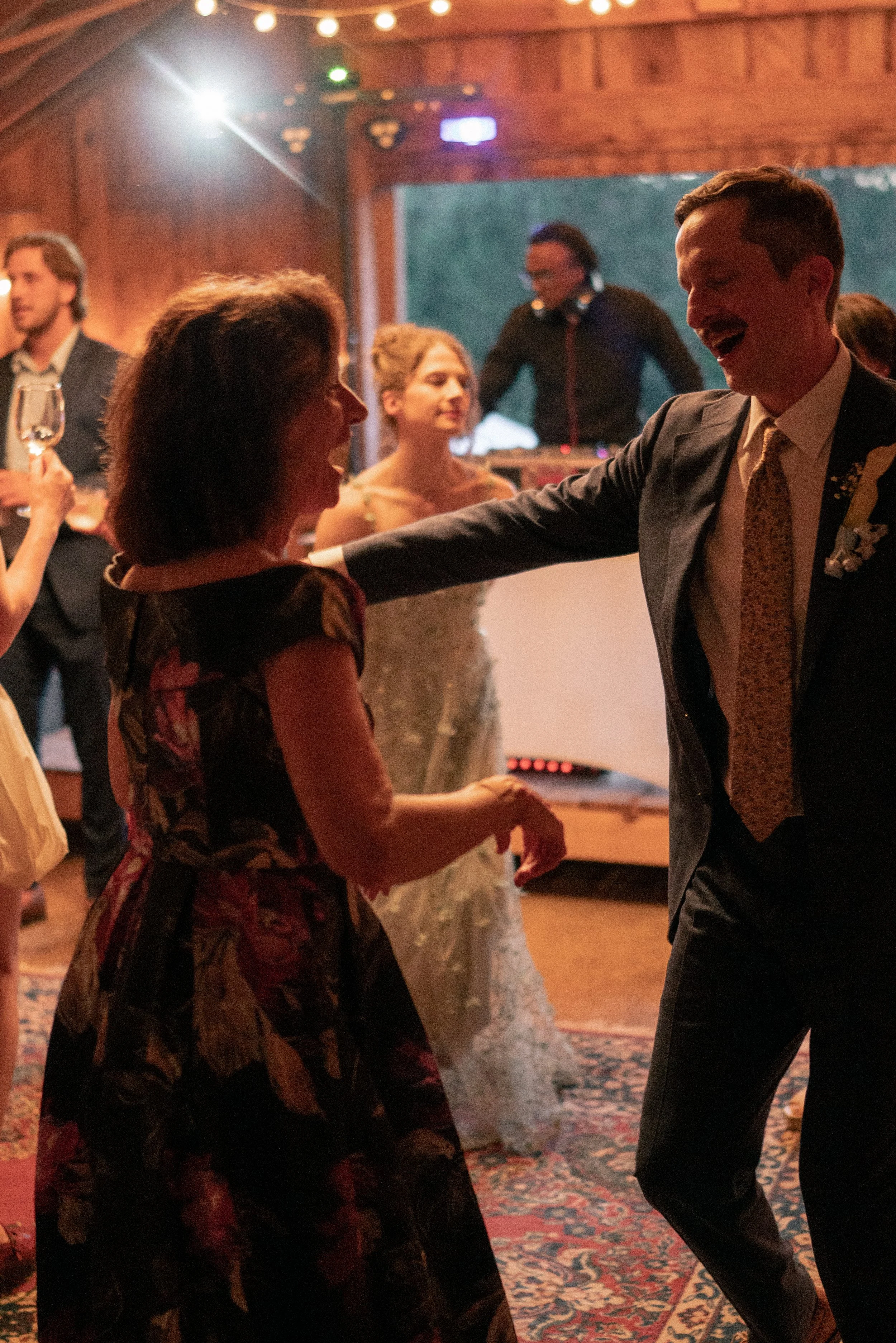 People dancing and socializing at a lively indoor event with wooden walls, string lights, and a DJ in the background.