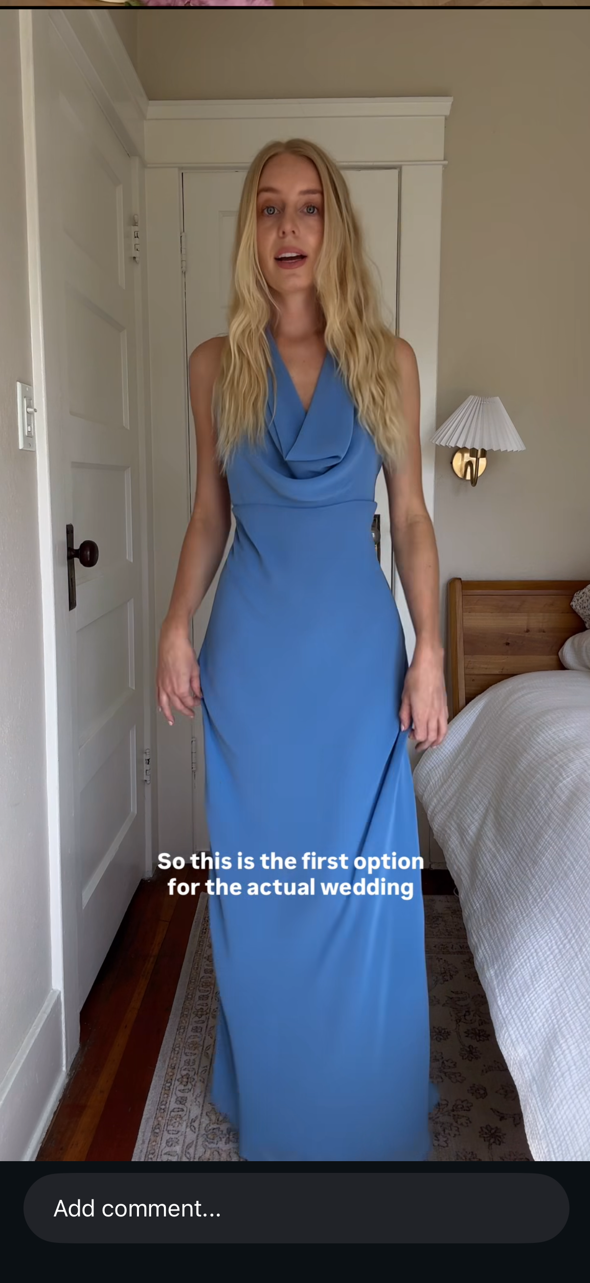 A woman with long blonde hair wearing a blue sleeveless dress standing in a bedroom, with a bed and wall-mounted lamp in the background. The text overlay reads, "So this is the first option for the actual wedding."