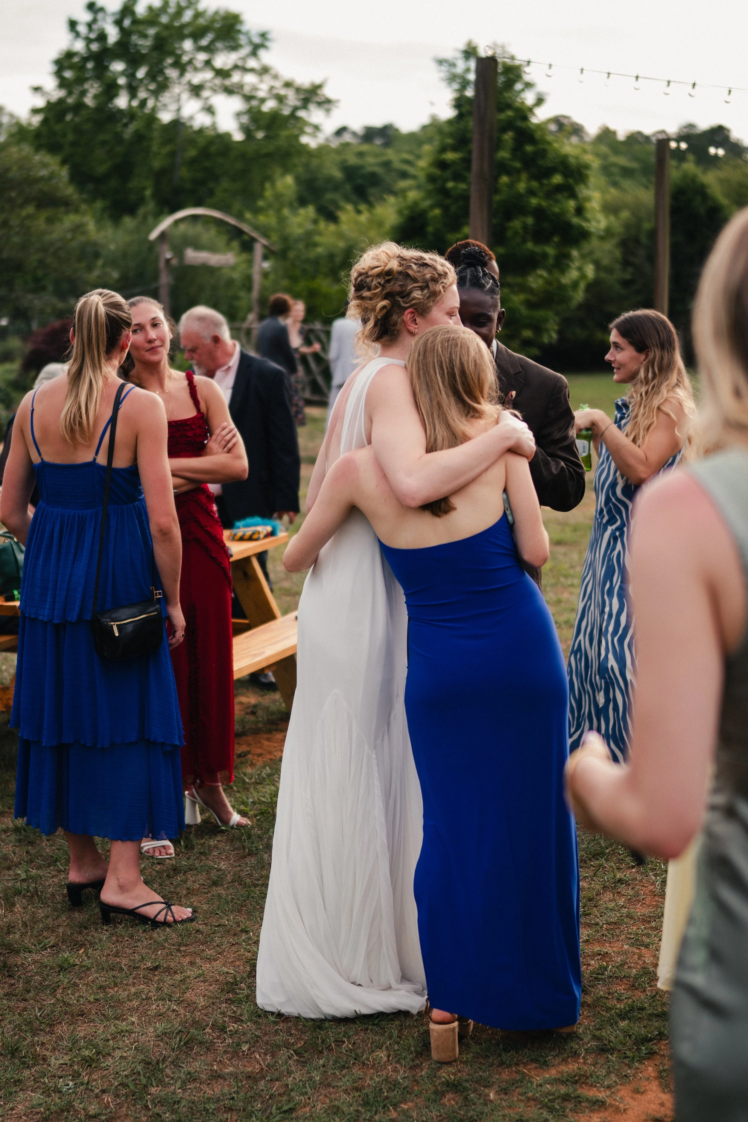 People gathered outdoors at a wedding reception, engaged in conversation, with trees, string lights, and a wooden structure in the background.
