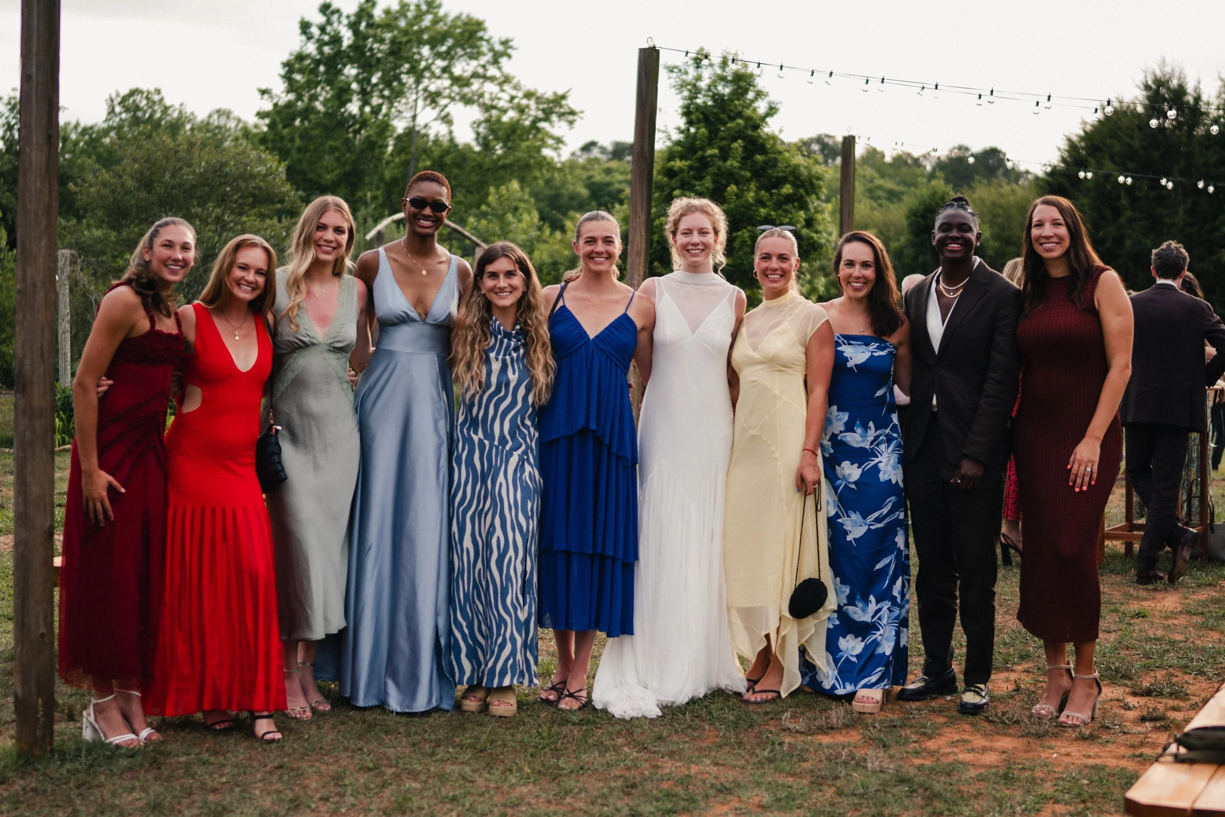 Group of women in colorful dresses standing outdoors, smiling for a photo, with string lights and trees in the background.