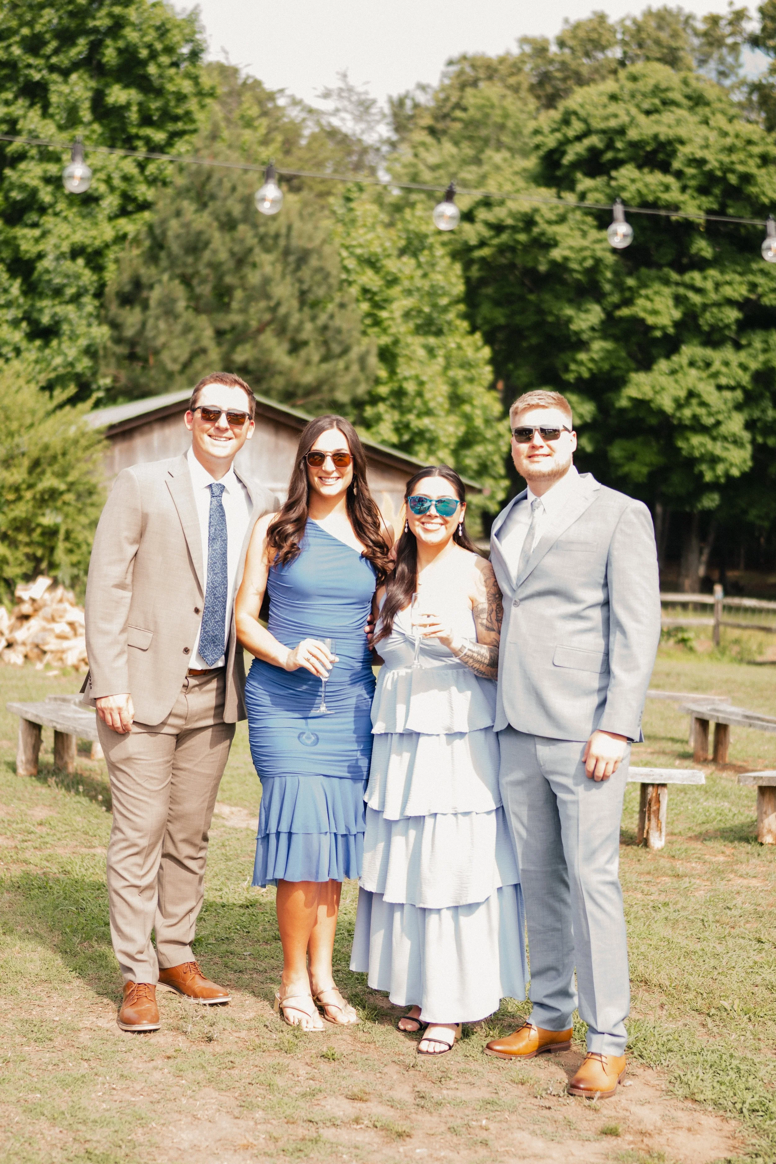Four people dressed in formal attire standing outside on a grassy area with trees and string lights overhead, smiling at the camera.